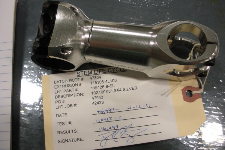 A metal bicycle stem resting on a reflective surface, accompanied by a printed tag detailing its specifications including batch number, extrusions, part number, description, and test results. The stem features a polished silver finish with black components.