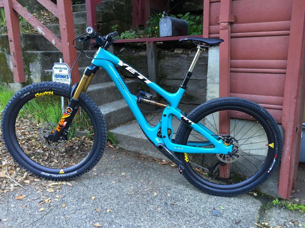 Yeti SB6: A bright blue mountain bike leaning against a stairway, with a sign in the background indicating home security. The bike features prominent suspension components and wide tires, set against a backdrop of greenery and a rustic building.