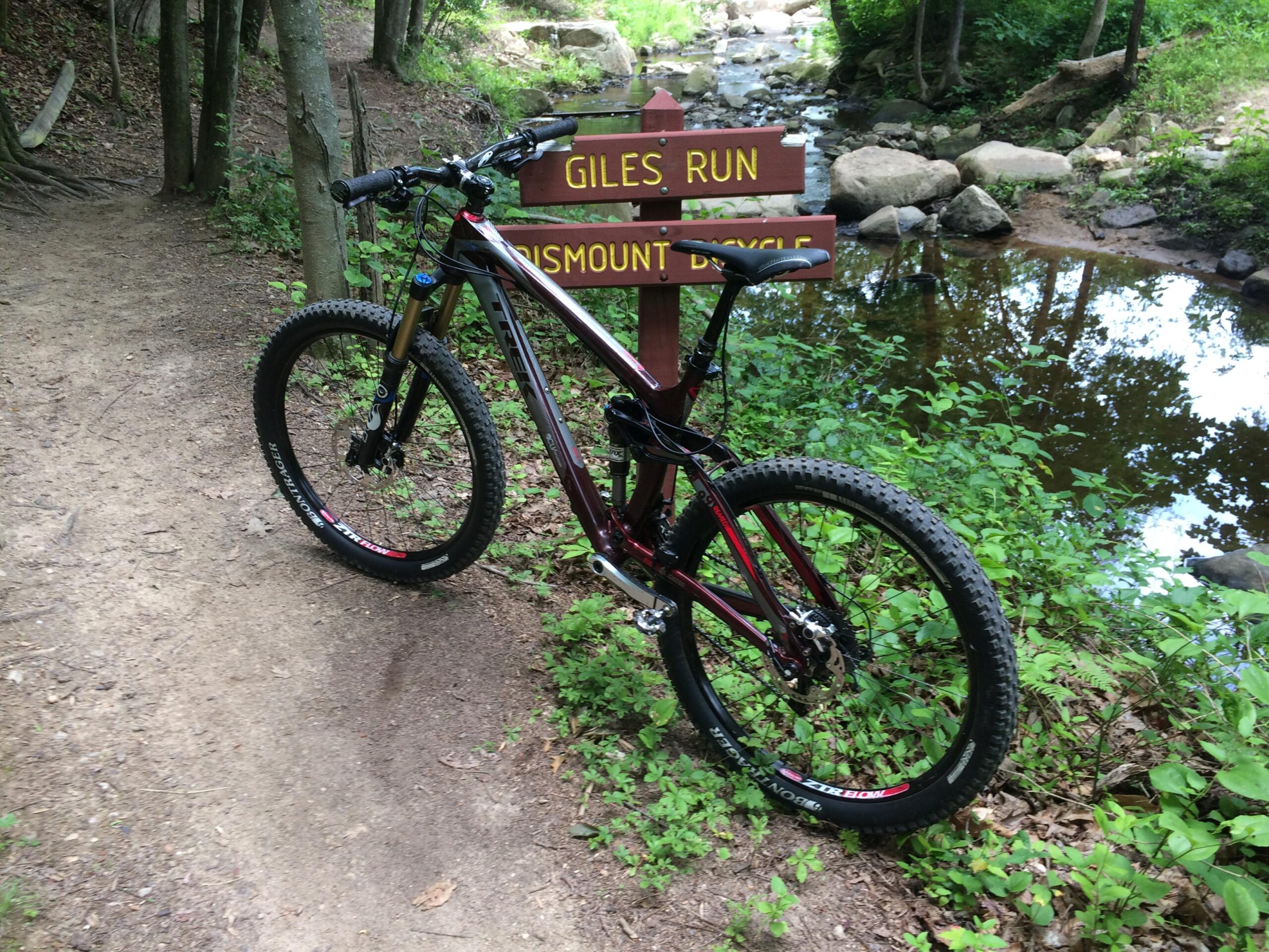 Trek Remedy 9.9: A mountain bike positioned near a trail sign that reads "Giles Run" and "Dismount Bicycle." The scene features a wooded area with a creek visible in the background, along with greenery and rocks. The bike's tires are designed for rugged terrains, and the path is made of dirt with patches of grass.