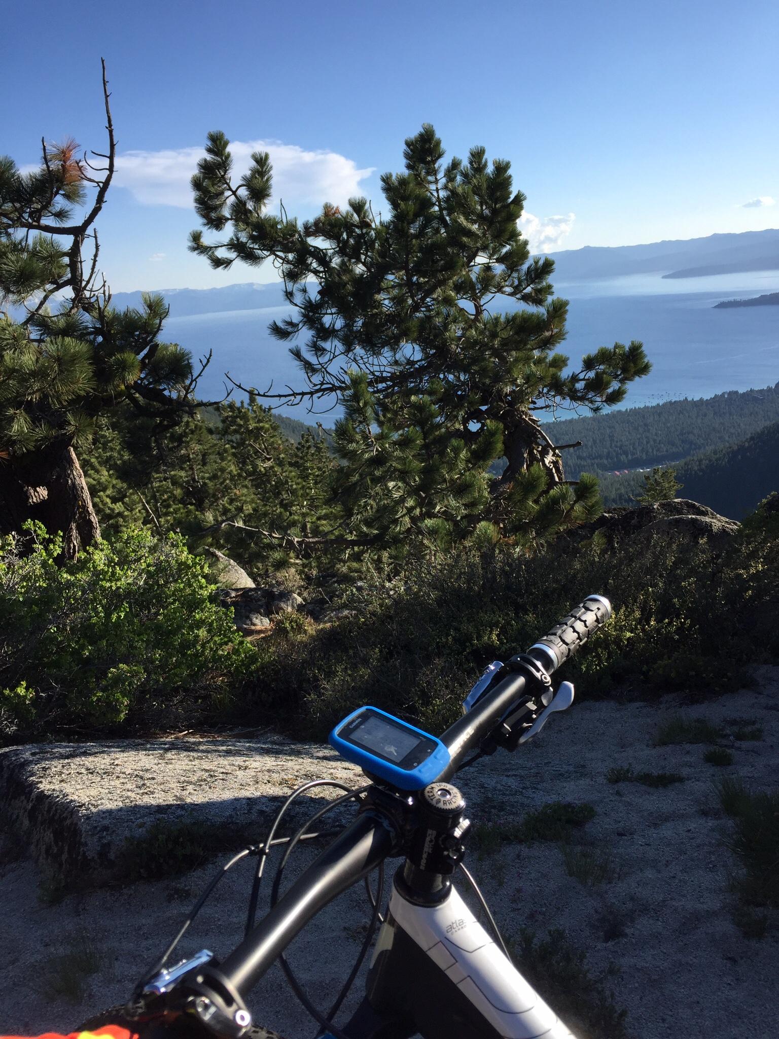 A mountain bike in the foreground with a digital display on the handlebars, overlooking a scenic view of a lake and forested mountains under a clear blue sky. Tahoe Rim Trail: Tahoe Meadows to Tunnel Creek Road / Flume Trail mountain bike trail.