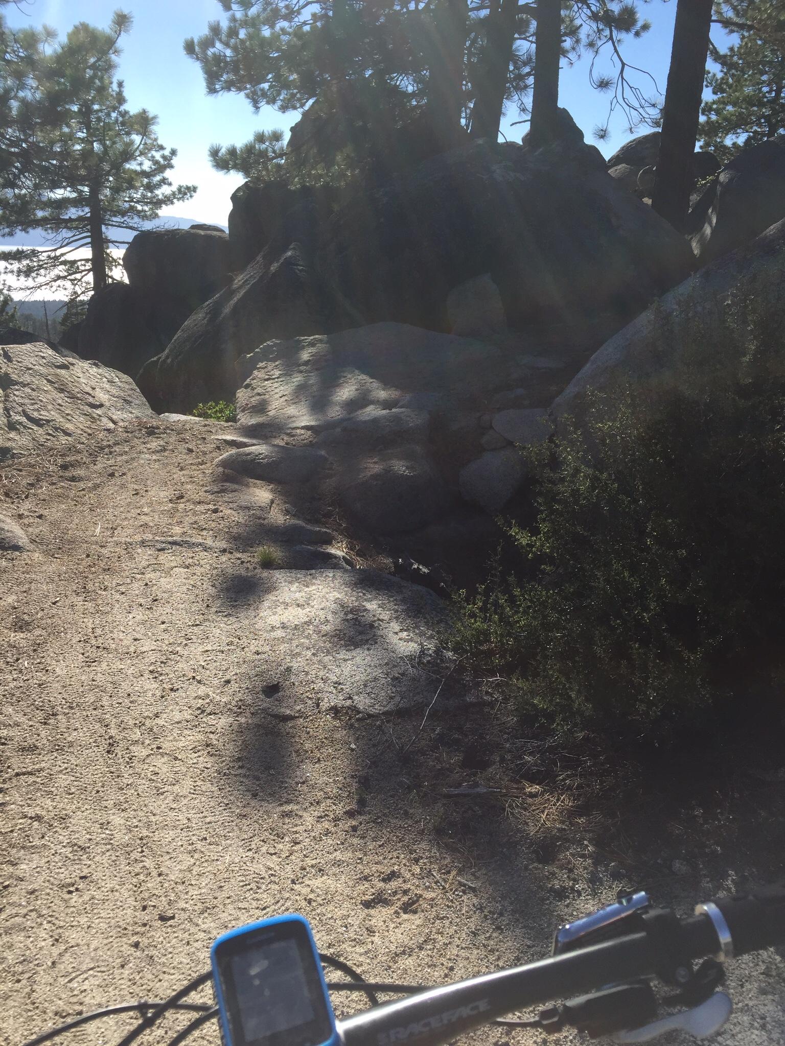 A rocky mountain biking trail surrounded by trees, with a clear blue sky in the background. The view shows the handlebars of a bike in the foreground, alongside a small digital device, indicating an outdoor adventure. Sunlight creates a warm glow, illuminating the path ahead. Tahoe Rim Trail: Spooner Summit to Kingsbury Grade mountain bike trail.