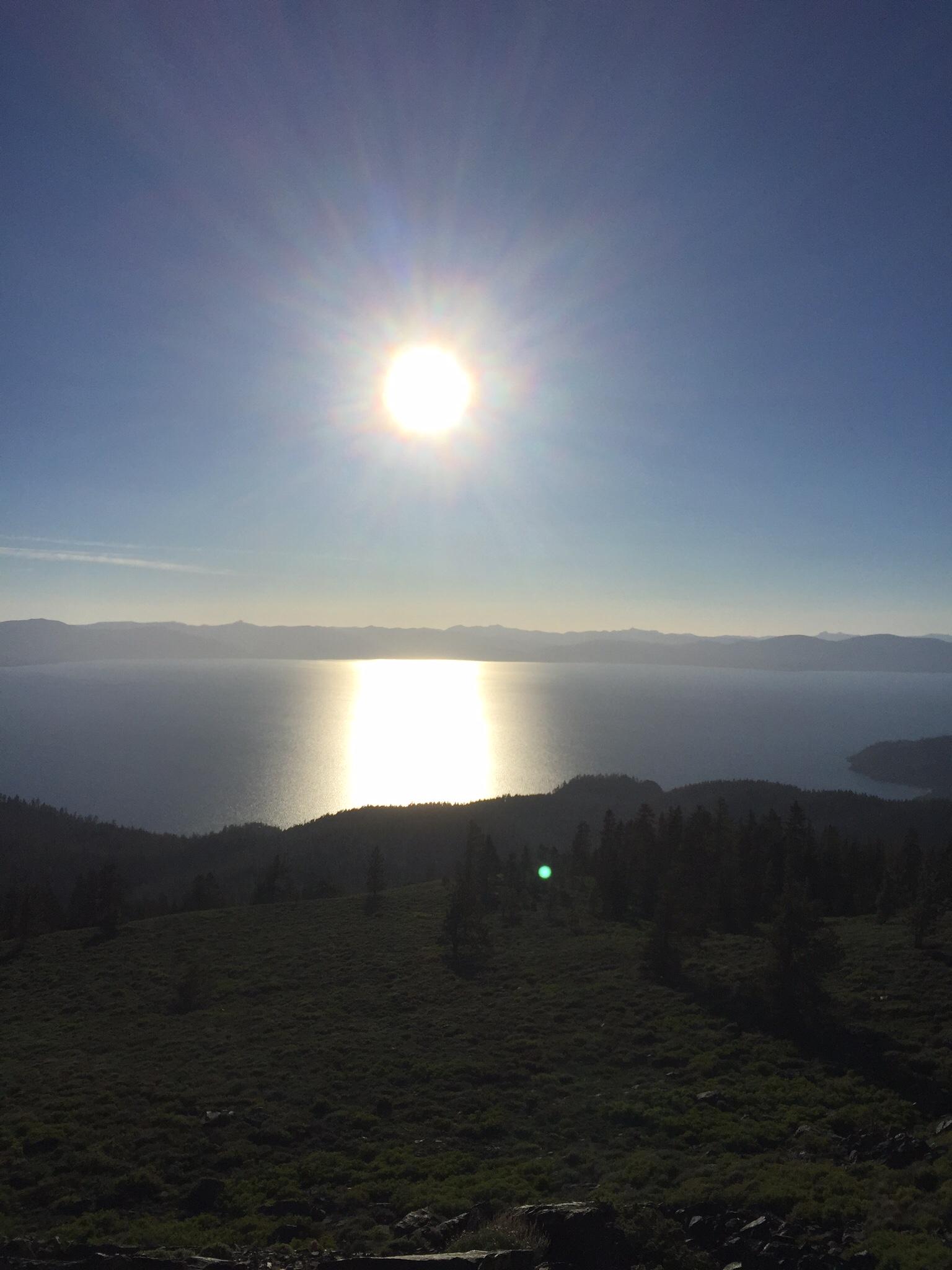 A bright sun shining in a clear blue sky over a tranquil lake, with sunlight reflecting on the water's surface. The foreground features lush green hills and scattered trees, while distant mountains outline the horizon. Tahoe Rim Trail: Spooner Summit to Kingsbury Grade mountain bike trail.