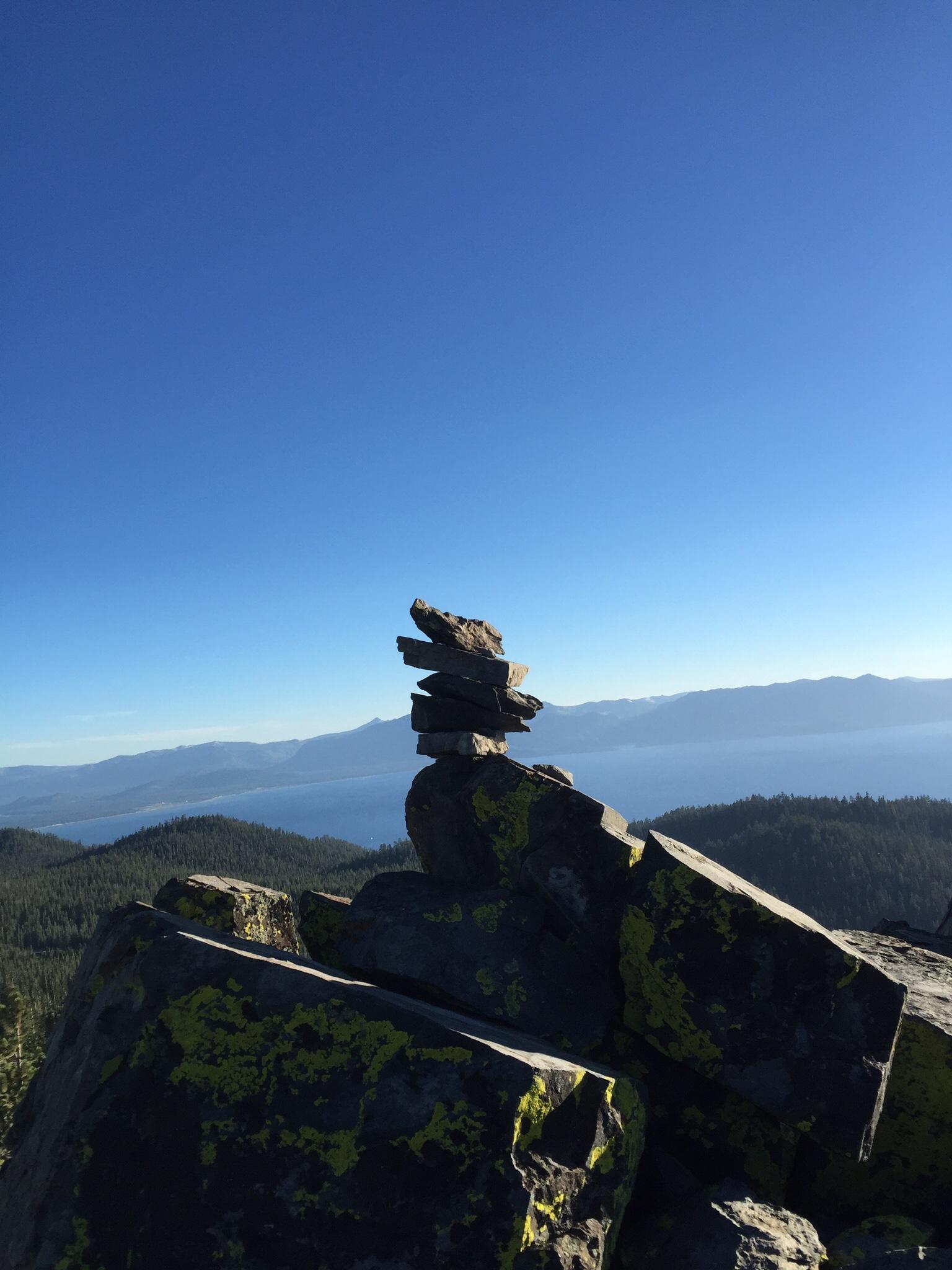 A stack of flat stones balanced on a pile of rocky boulders, set against a clear blue sky and a scenic mountain landscape with a lake in the background. The boulders are covered with yellow-green lichen, adding color to the natural setting. Tahoe Rim Trail: Spooner Summit to Kingsbury Grade mountain bike trail.