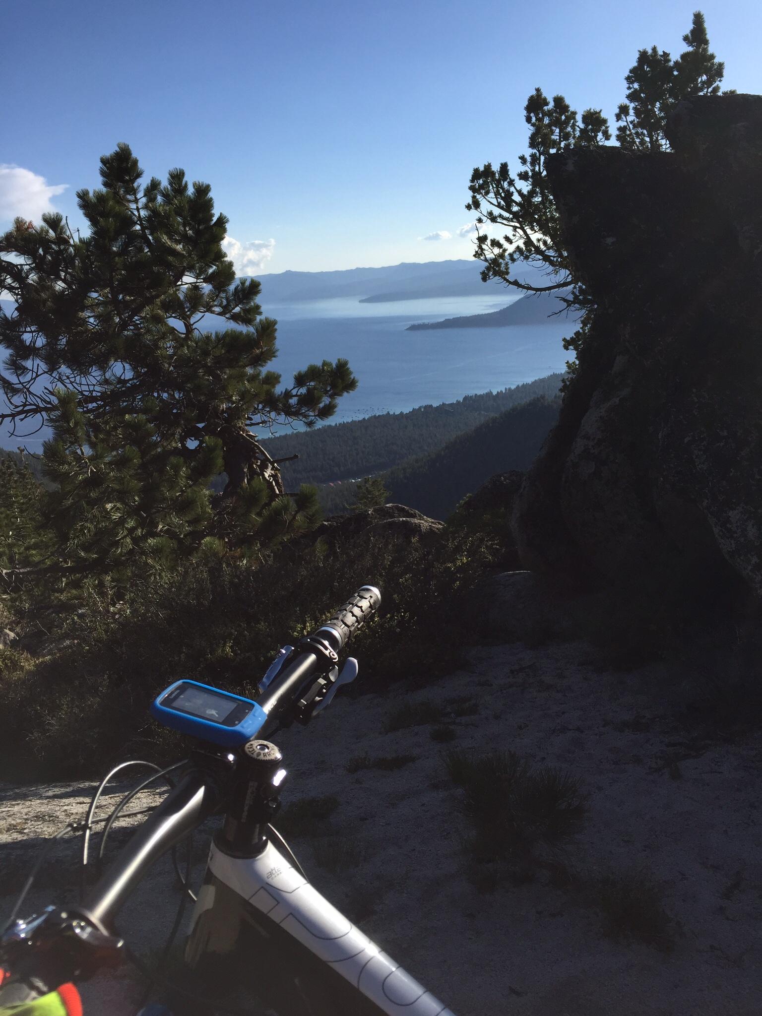 A mountain bike handlebar is in the foreground, equipped with a GPS device, while the background showcases a stunning view of a lake surrounded by mountains and forested hills under a clear blue sky. Tahoe Rim Trail: Tahoe Meadows to Tunnel Creek Road / Flume Trail mountain bike trail.