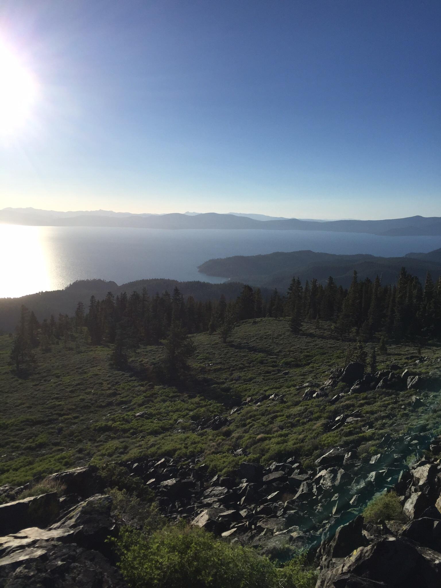 A panoramic view of a mountainous landscape featuring a serene lake, with sunlight illuminating the scene. The foreground includes rocky terrain and lush greenery, while distant mountains are visible on the horizon under a clear blue sky. Tahoe Rim Trail: Spooner Summit to Kingsbury Grade mountain bike trail.