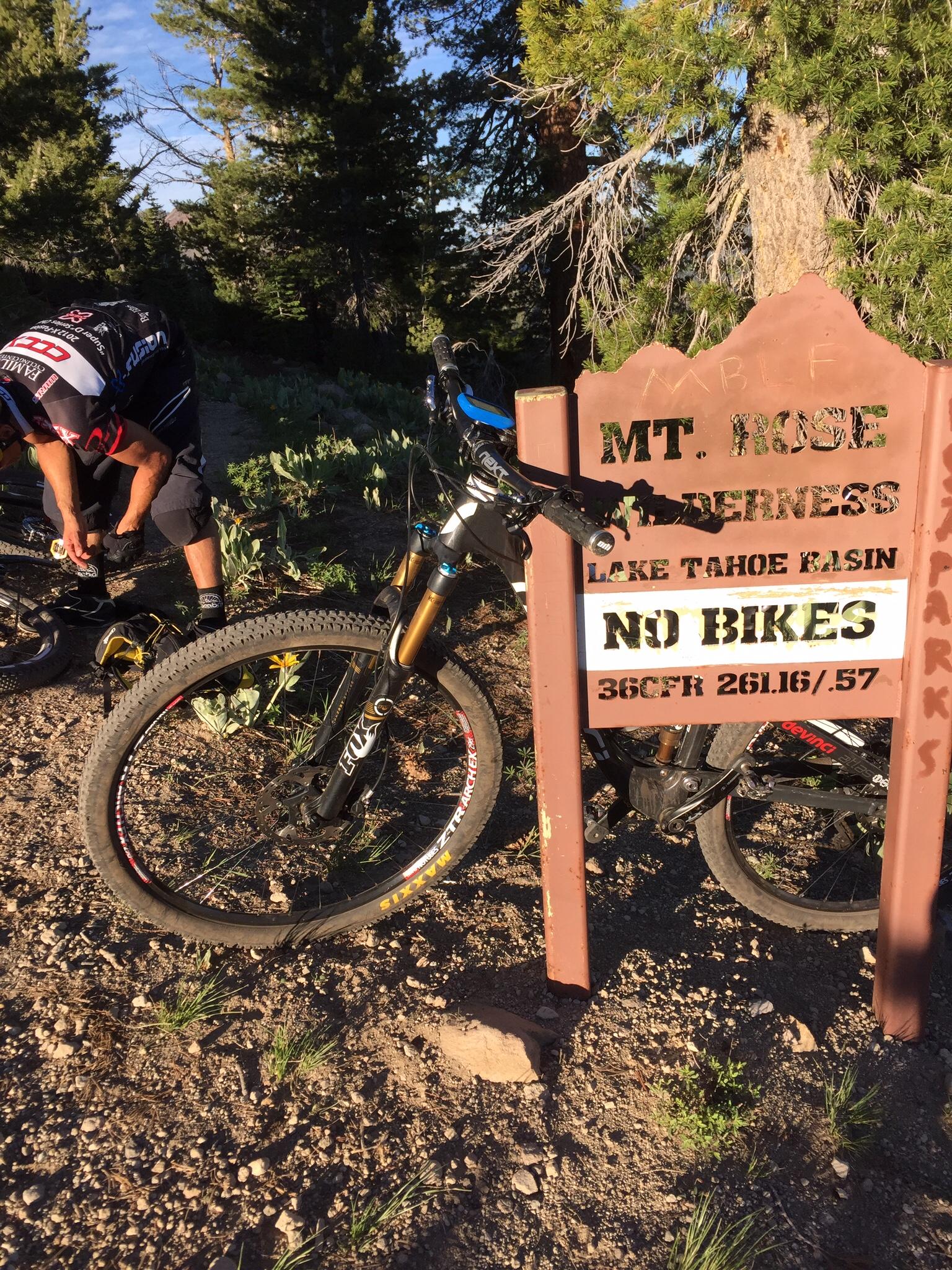 A mountain biker adjusts their gear near a sign indicating the Mt. Rose Wilderness area in the Lake Tahoe Basin, with a clear message reading "No Bikes." A mountain bike is resting close to the sign, surrounded by natural vegetation and trees. The sun casts a warm light on the scene, suggesting a clear, serene day. Tahoe Rim Trail: Brockway to Watson Lake mountain bike trail.