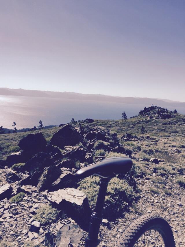 A scenic view of a mountainous landscape with a clear blue lake in the distance, featuring rocky terrain and patches of greenery. In the foreground, the seat and part of the frame of a mountain bike are visible, indicating an outdoor adventure setting. Tahoe Rim Trail: Spooner Summit to Kingsbury Grade mountain bike trail.