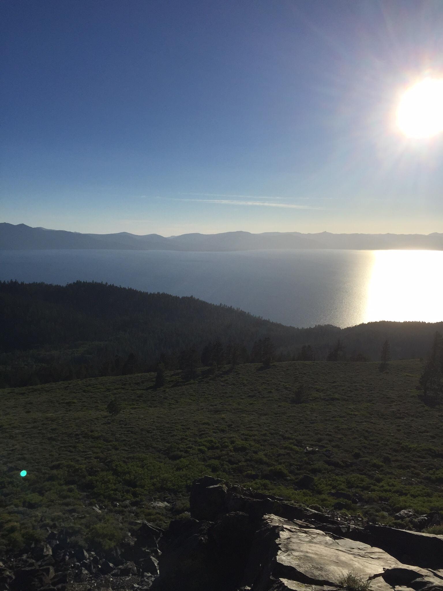 A serene landscape view showcasing a vast lake surrounded by rolling hills under a clear blue sky. The sun is shining brightly in the top right corner, reflecting off the water's surface and illuminating the greenery in the foreground. Tahoe Rim Trail: Spooner Summit to Kingsbury Grade mountain bike trail.