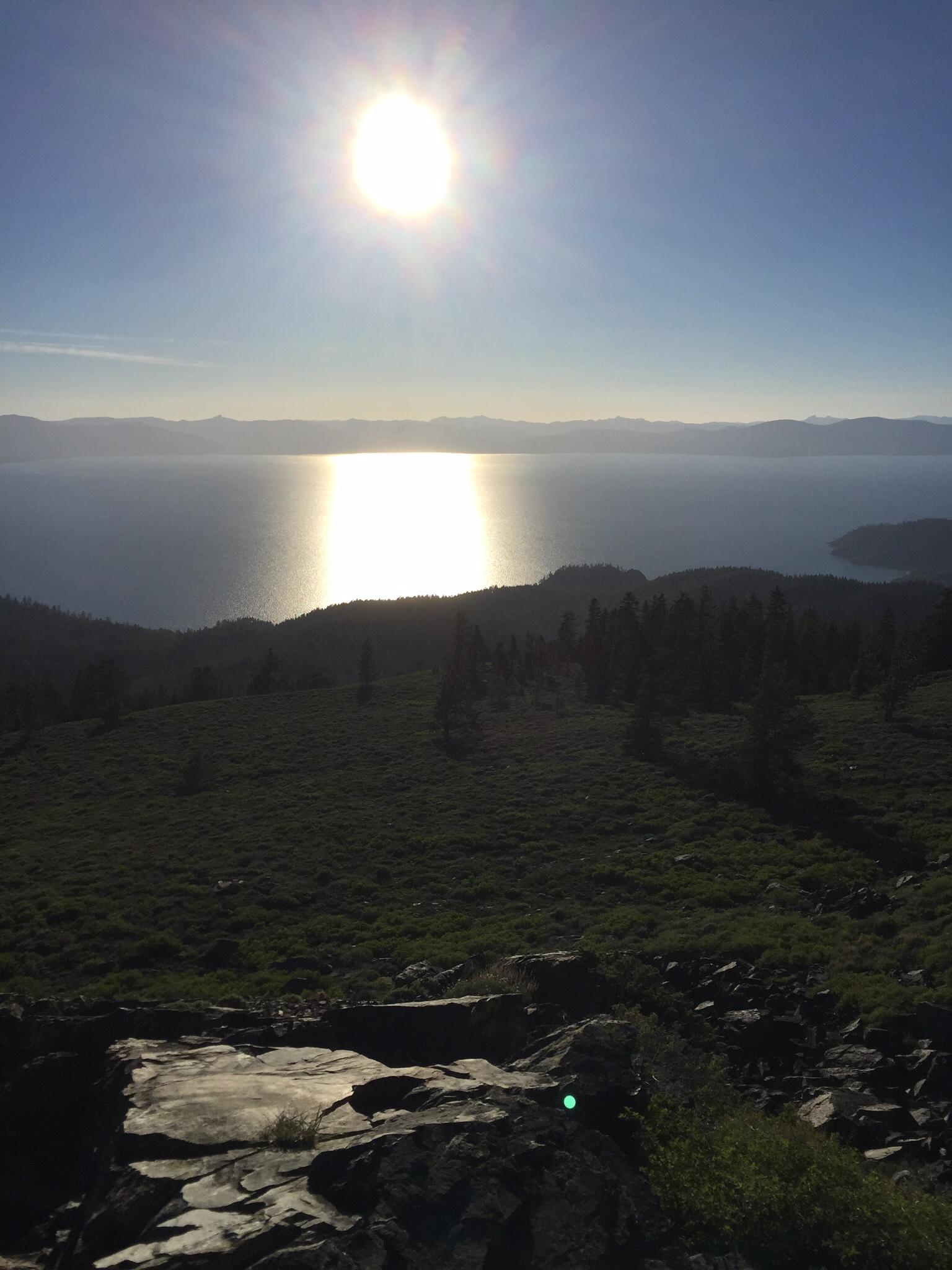 Sun shining over a tranquil lake, reflecting light on the water's surface, with lush green hillside and rocky foreground in a serene outdoor landscape. Blue sky and distant mountains create a peaceful backdrop. Tahoe Rim Trail: Spooner Summit to Kingsbury Grade mountain bike trail.