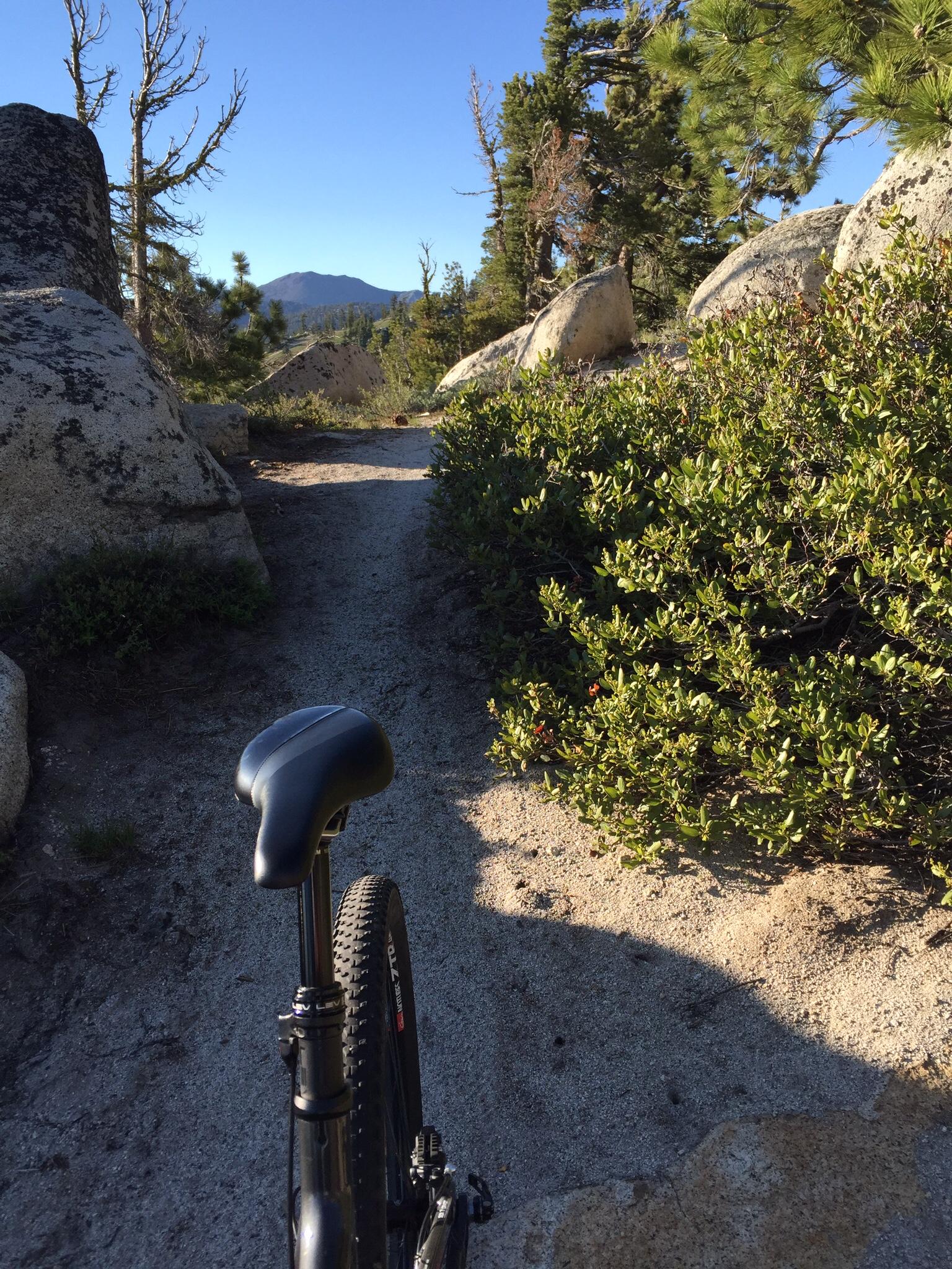 A mountain bike resting on a sandy trail surrounded by large boulders and greenery, with a clear blue sky and mountains visible in the distance. Tahoe Rim Trail: Tahoe Meadows to Tunnel Creek Road / Flume Trail mountain bike trail.