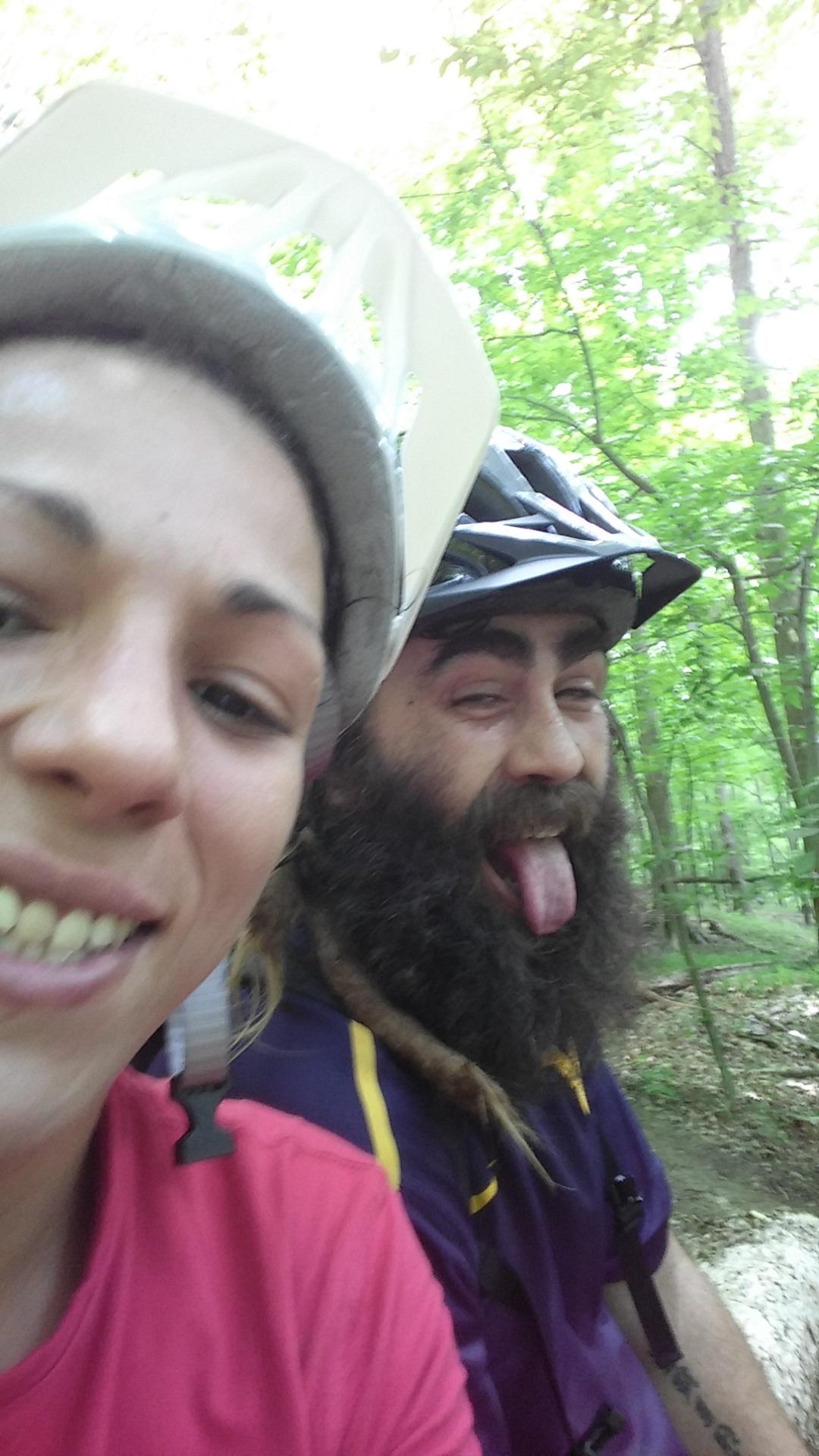 A close-up selfie of two people wearing bike helmets, smiling at the camera. The background is a lush green forest with trees. The person on the left has long hair and is wearing a red t-shirt, while the person on the right has a long beard and is sticking out their tongue playfully. Mountwood mountain bike trail.
