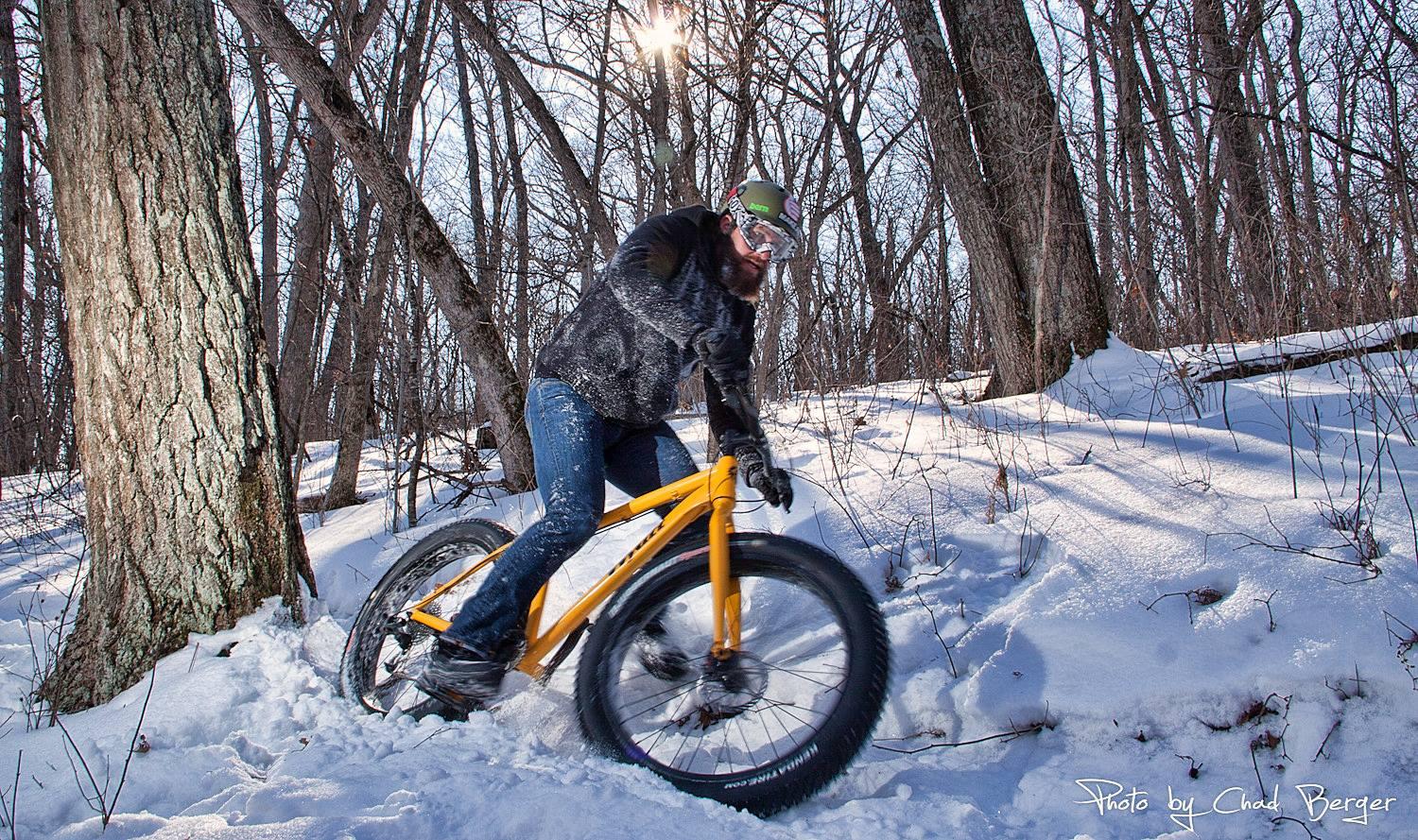 A person riding a bright yellow fat bike through snowy terrain in a wooded area, with trees and sunlight filtering through branches in the background. The rider is wearing a helmet and goggles, and snow is being kicked up from the bike