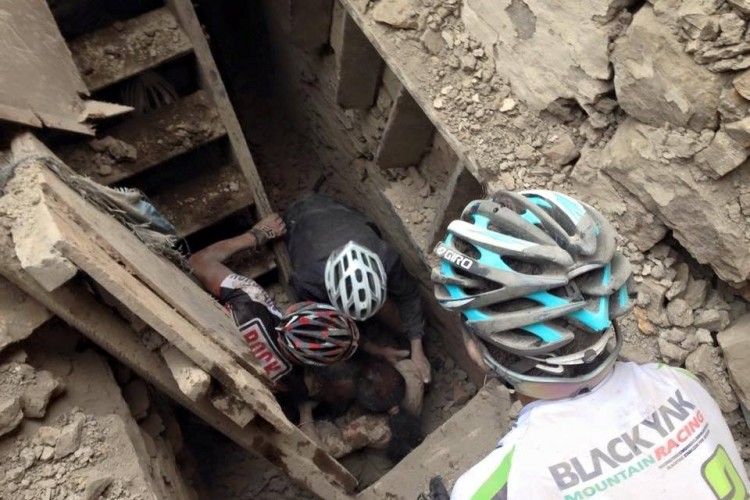 A group of mountain bikers assisting a person trapped in a narrow, debris-filled space, likely a collapsed structure. The scene shows dust and rubble around, and the bikers are focused on helping the individual while wearing helmets.