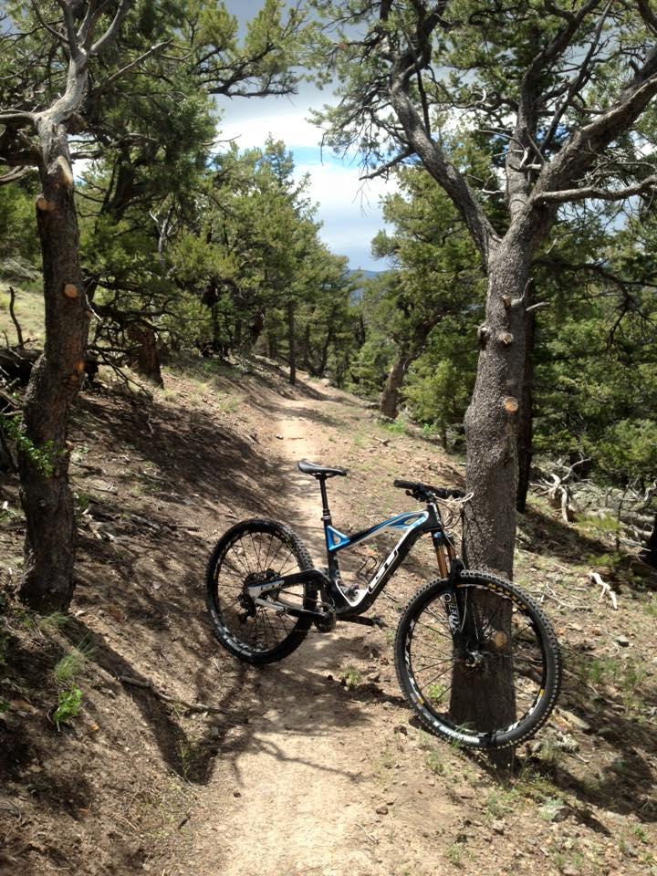 A mountain bike resting on a dirt trail surrounded by trees in a scenic outdoor setting. The path is narrow, winding through greenery, leading into the distance. Bright blue skies peek through the tree branches, indicating a clear day. Cottonwood mountain bike trail.