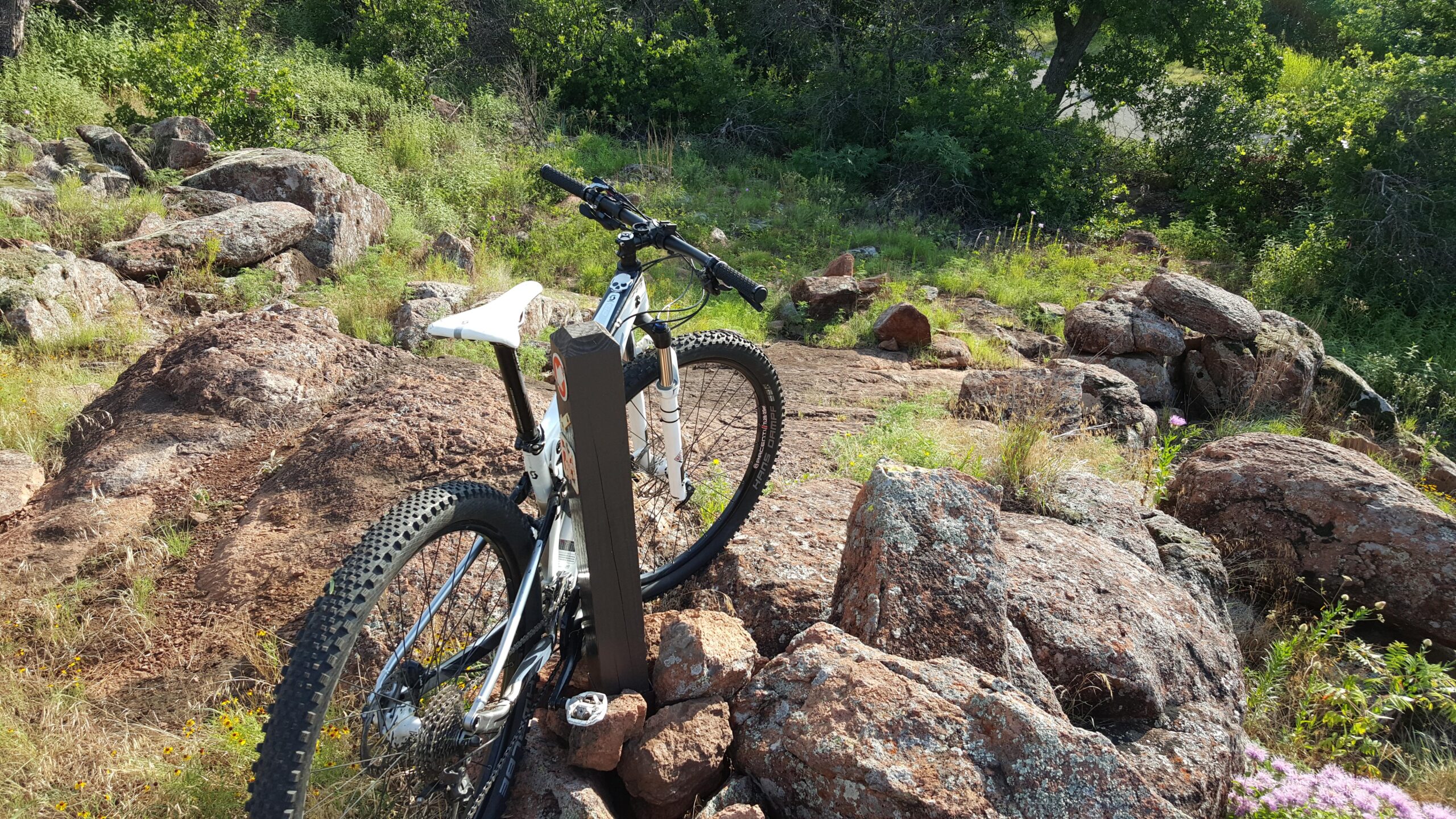 A mountain bike parked on rocky terrain surrounded by grassy areas and shrubs. The scene features large boulders and vibrant greenery, suggesting an outdoor trail suitable for biking adventures. Lake Lawtonka Trails mountain bike trail.
