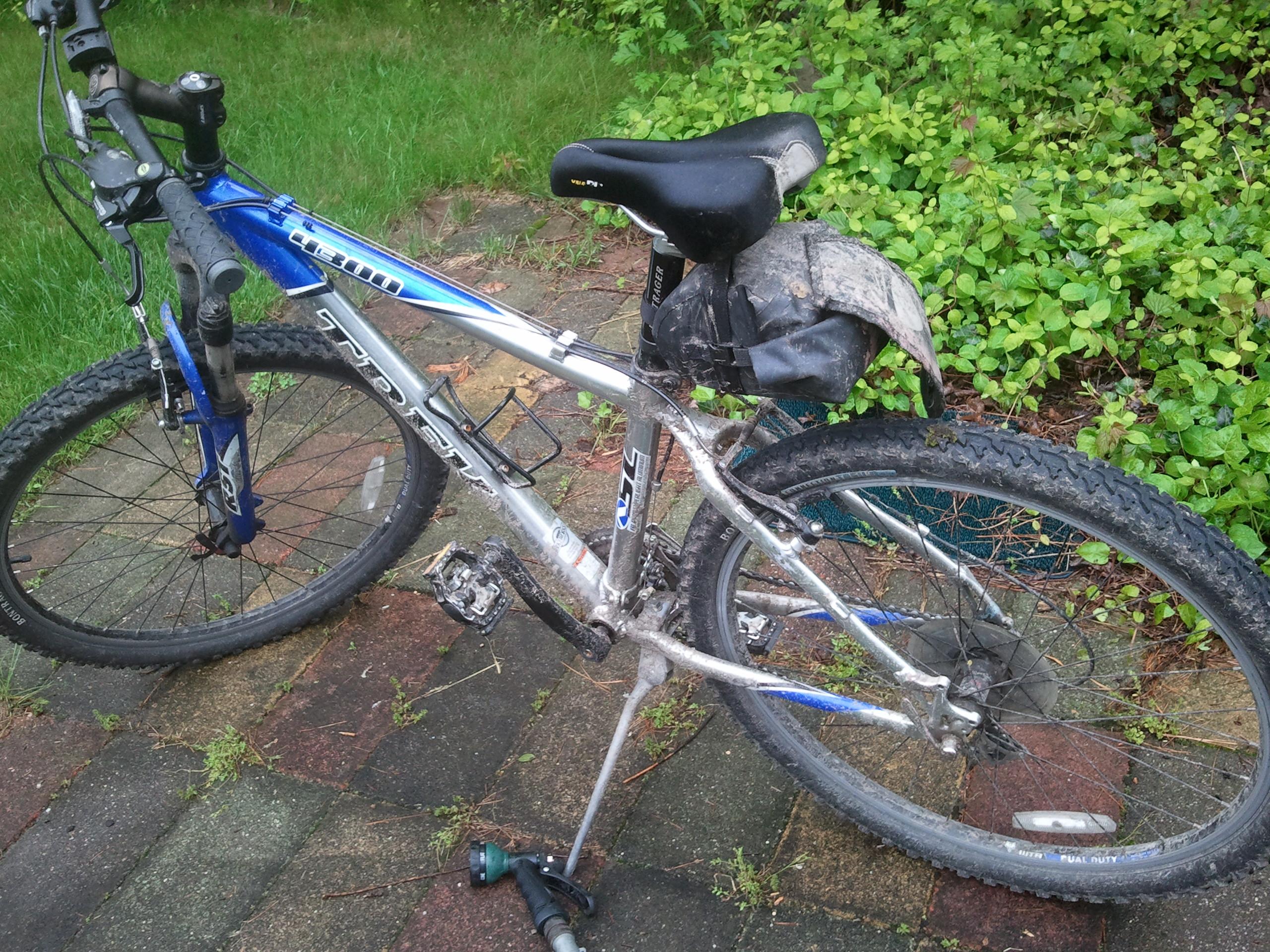 Trek 4300: A close-up view of a silver and blue mountain bike resting on a brick pathway, featuring a dirty frame and muddy tires. A small black saddlebag is attached to the back of the bike. In the background, there is a patch of grass and green foliage.