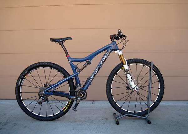 Santa Cruz Tallboy Carbon: A blue mountain bicycle with a lightweight frame and thick tires, positioned on a bike stand against a neutral background. The bike features front suspension and has distinct branding visible on the frame.