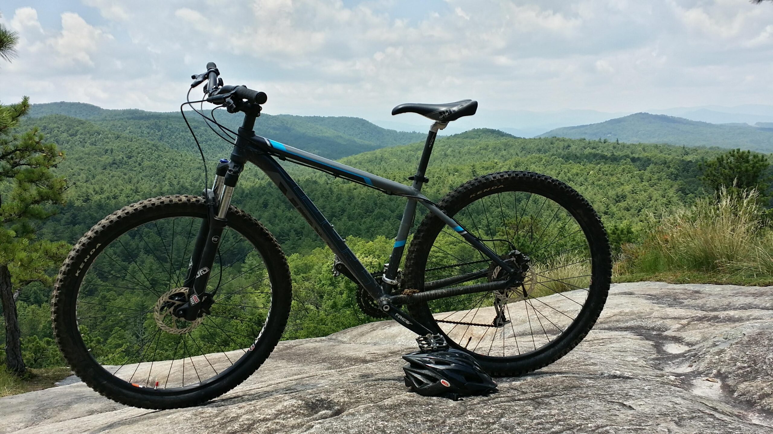 Gary Fisher Mamba: A mountain bike rests on a rocky ledge, overlooking a lush green landscape of rolling hills and trees under a partly cloudy sky. A black bicycle helmet is placed on the ground next to the bike.