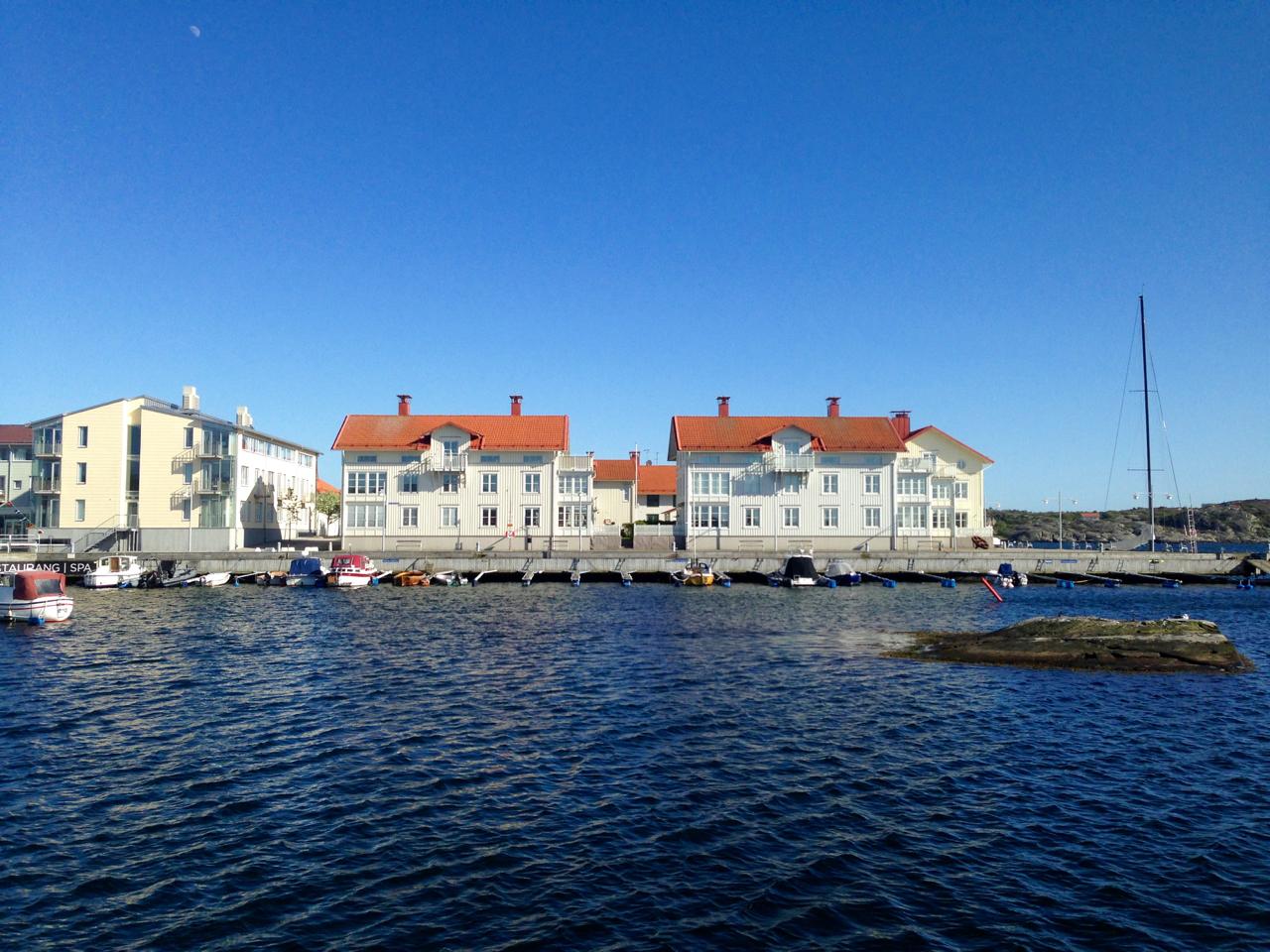 A waterfront scene featuring two buildings with red rooftops and white façades, located beside a calm blue body of water. Small boats are moored along a pier, and a sailboat is visible in the background against a clear blue sky. Marstrand Island Trail System mountain bike trail.