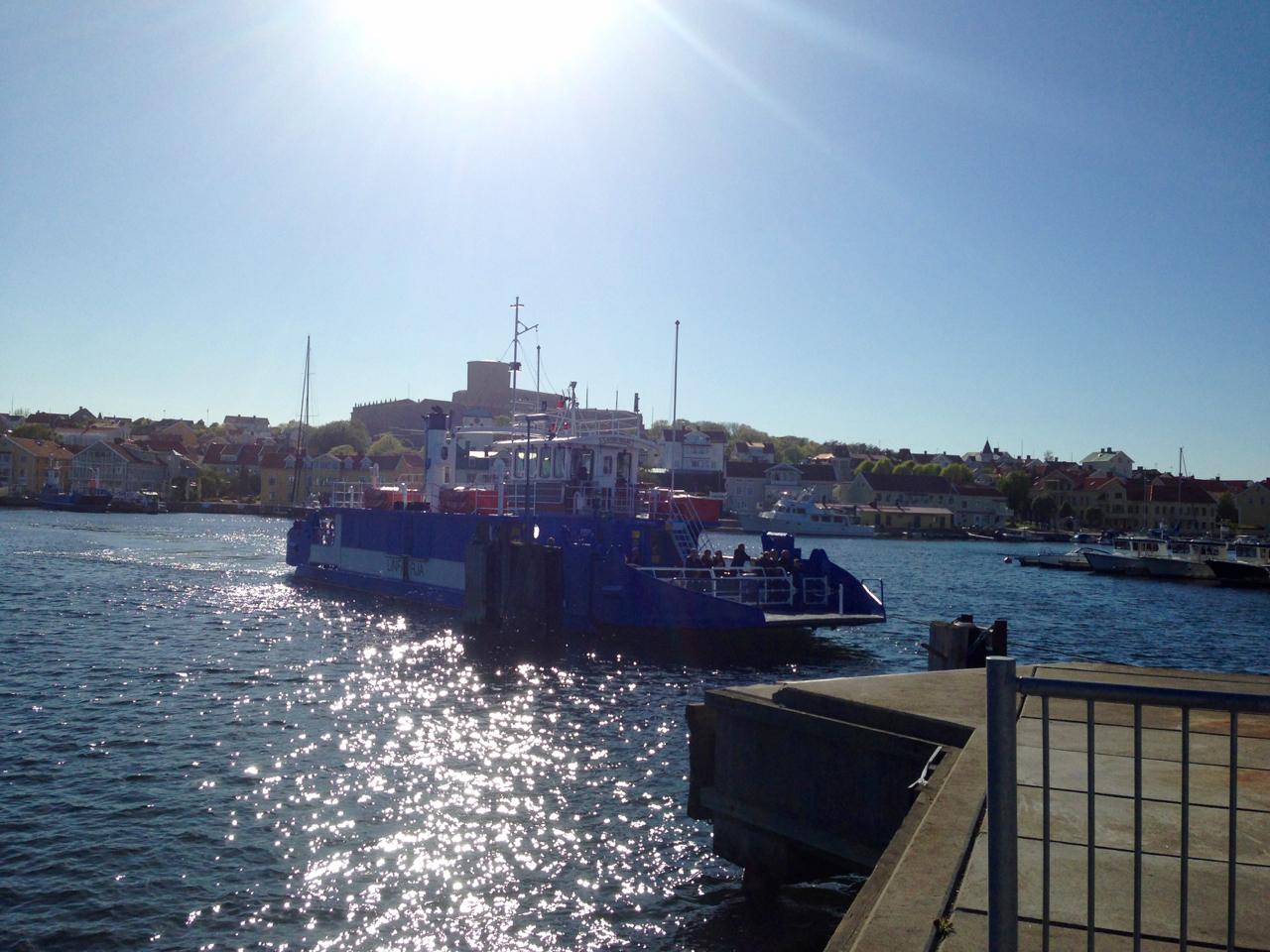 A blue ferry boat is docked at a pier on a sunny day, with the sun shining brightly in the sky. The water sparkles in the sunlight, and colorful buildings can be seen lining the waterfront in the background. Several boats are moored nearby, and a clear blue sky completes the picturesque scene. Marstrand Island Trail System mountain bike trail.