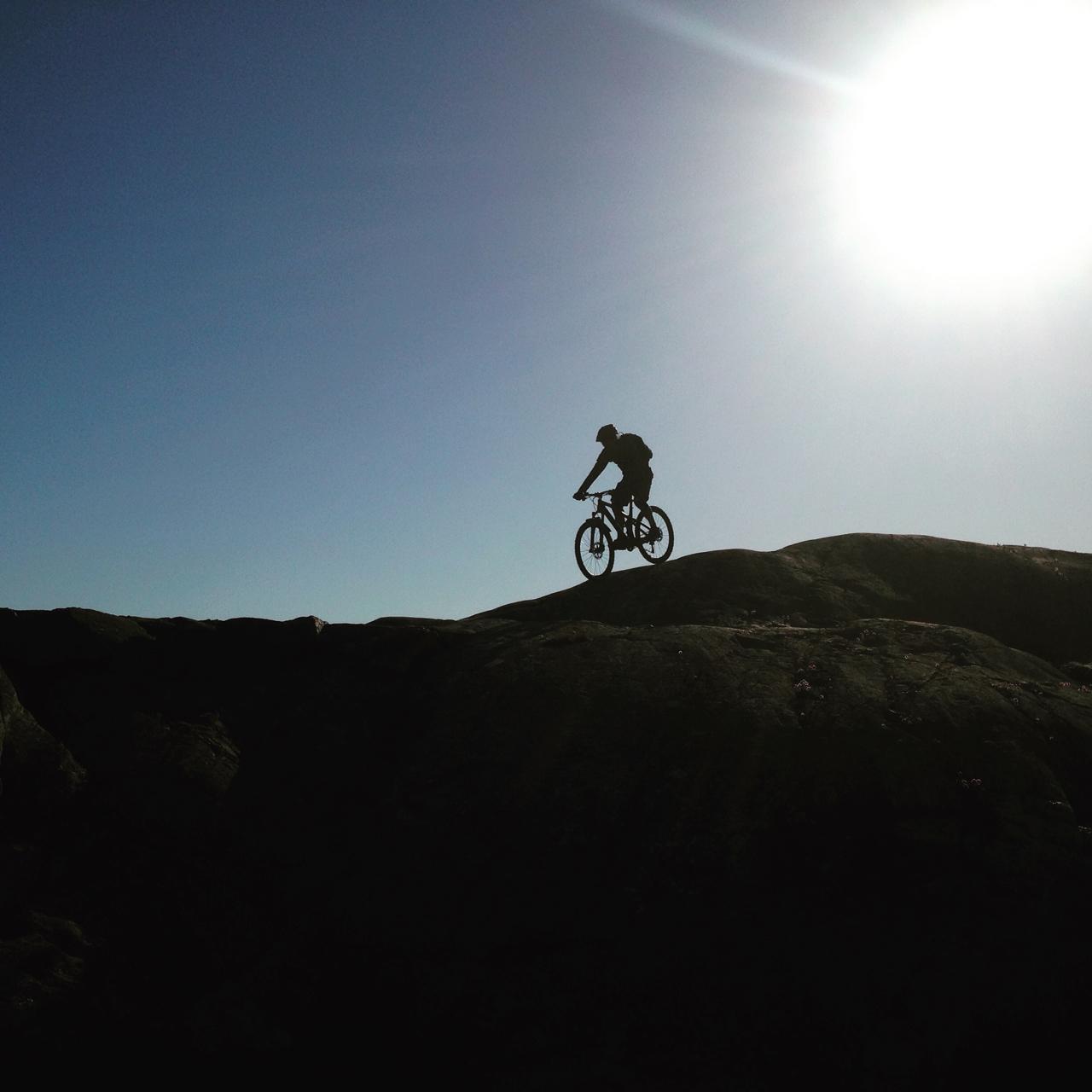 Silhouette of a mountain biker ascending a rocky terrain under a clear blue sky, with the sun shining brightly in the corner of the image. Marstrand Island Trail System mountain bike trail.