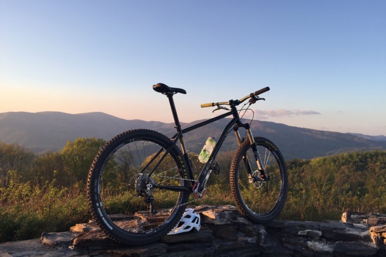 A black mountain bike rests on a stone wall, overlooking a scenic view of rolling hills and mountains at sunset. The sky is a gradient of colors, transitioning from golden to soft blue, with a few clouds scattered. The bike has chunky tires and a water bottle attached, indicating readiness for an outdoor adventure.