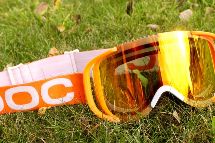 An orange ski goggles resting on green grass, featuring a white adjustable strap with the brand name prominently displayed. The lenses are reflective, showcasing a golden hue.