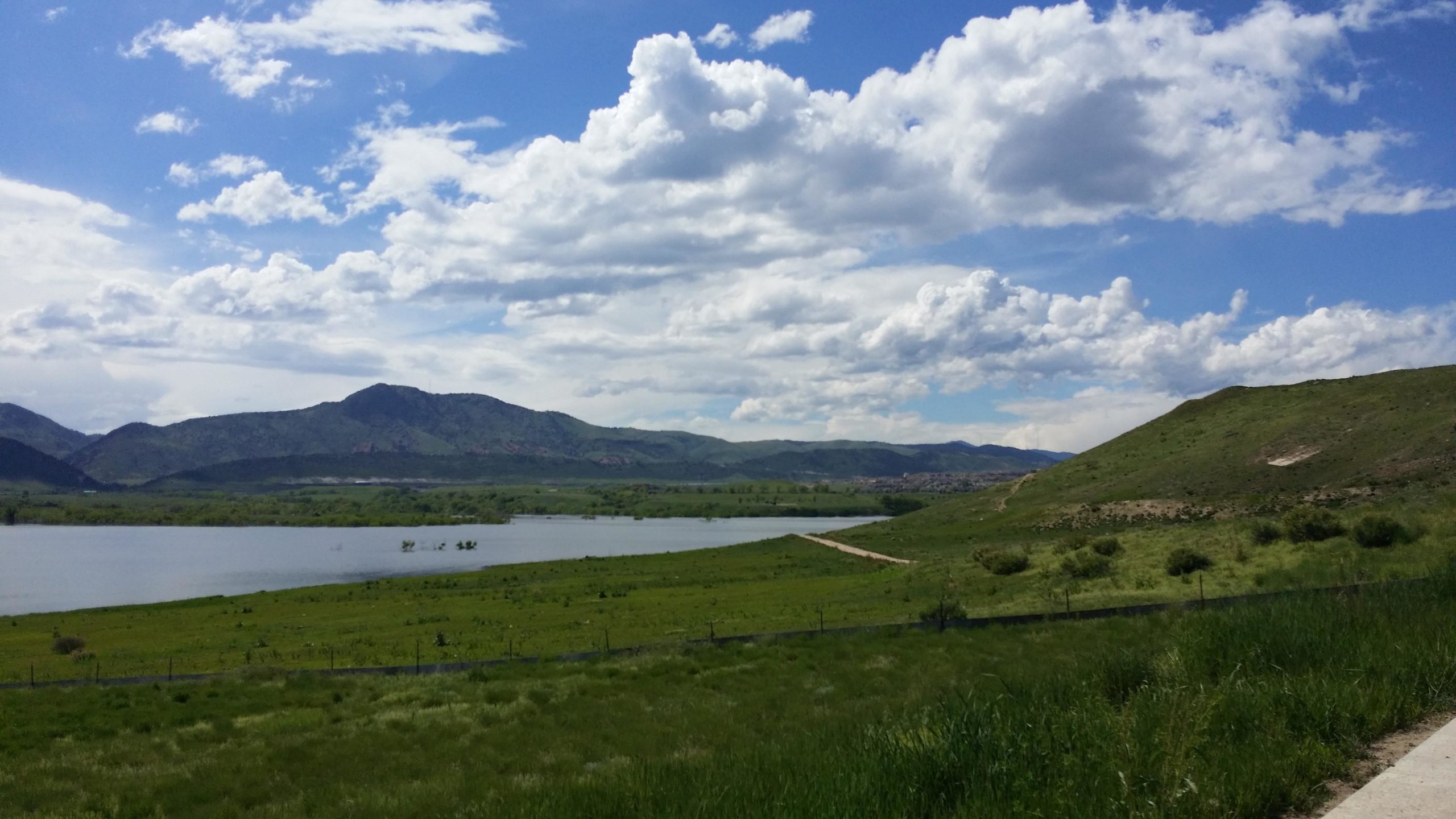 A scenic landscape featuring a calm lake surrounded by lush greenery, with rolling hills and mountains in the background under a partly cloudy blue sky. A dirt path winds along the shoreline, inviting exploration of the serene natural environment. Bear Creek Lake Park mountain bike trail.