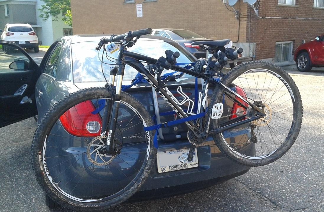 Specialized CRAVE 29: A black mountain bike secured to the rear of a gray car using a bike rack. The bike is dirty, indicating recent use, and is held in place with blue straps. The car's back is partially visible, showing a license plate and a glimpse of the interior through an open door. In the background, other vehicles and buildings can be seen.