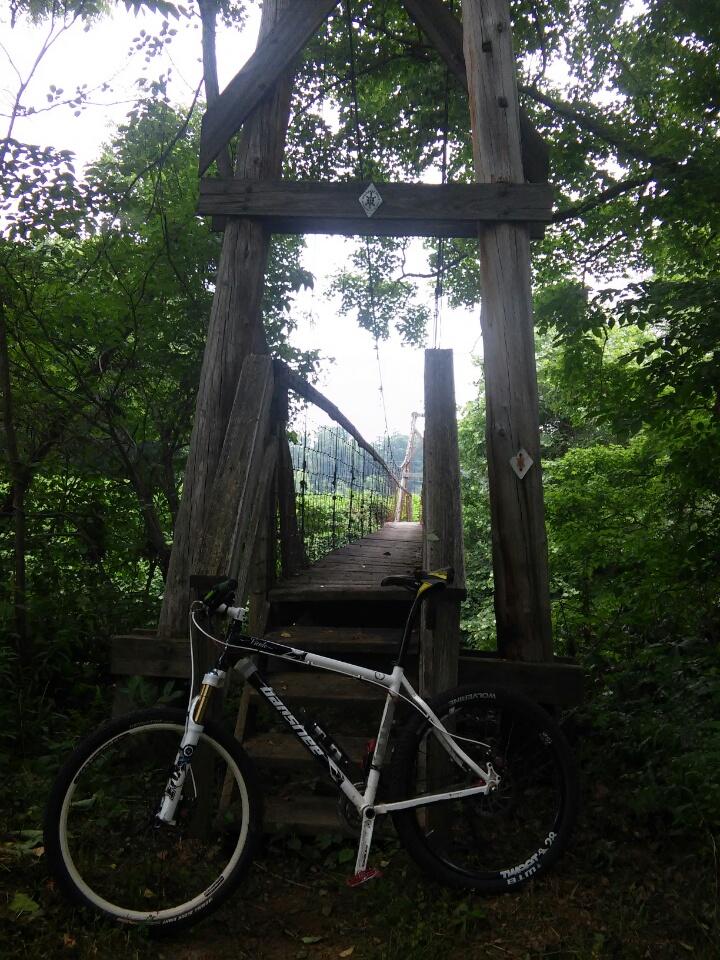 A mountain bike rests on the ground beside a wooden suspension bridge surrounded by lush greenery. The bridge, made of rustic wooden beams, leads through a dense forest, with trees framing the scene and a faint path visible in the background. The sky is overcast, hinting at a peaceful, secluded nature setting. Cave-run Lake mountain bike trail.