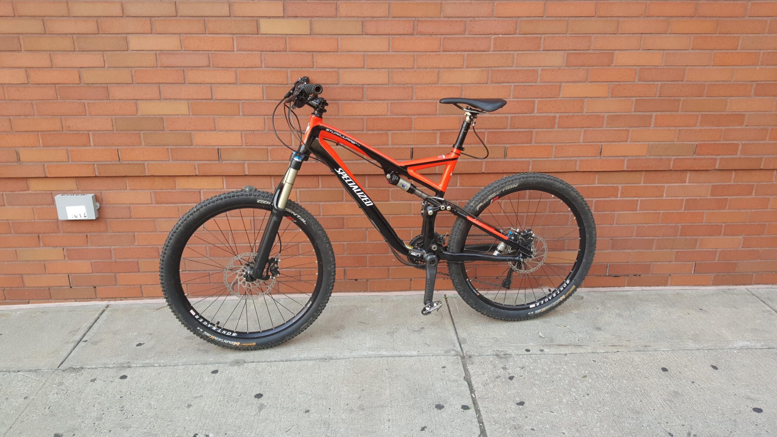 Specialized Stumpjumper FSR Elite: A black and orange mountain bike leaning against a brick wall, featuring thick tires and a suspension system suitable for off-road terrain.