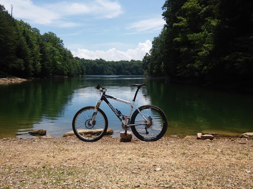 A mountain bike positioned on a rocky lakeshore, with calm green waters reflecting the blue sky and white clouds. Lush trees line the landscape in the background, creating a serene nature setting. Cane Creek (sheltowee Trace Trail) mountain bike trail.