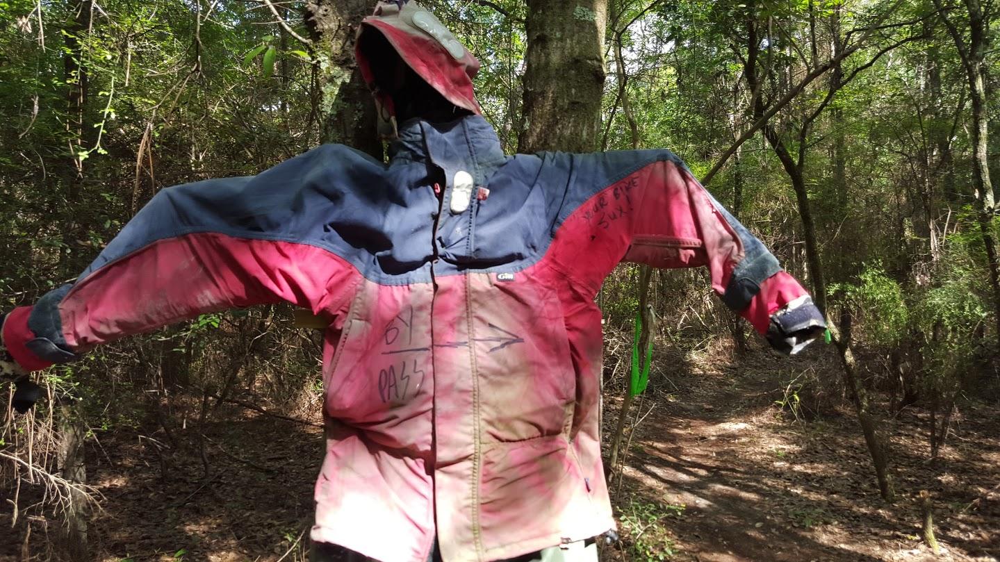 A colorful jacket hanging from a tree in a wooded area, with visible dirt stains and handwritten messages on the fabric. The background features dense greenery and a dirt path. Games Loop mountain bike trail.