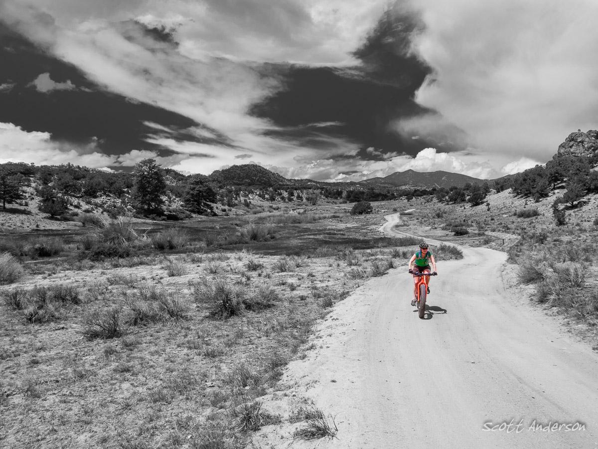 A person riding a mountain bike on a dirt trail through a scenic landscape, featuring rolling hills and sparse vegetation. The image is primarily in black and white, with the cyclist