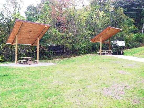 Two wooden pavilions with slanted roofs are positioned on a grassy area, surrounded by trees with changing foliage. Each pavilion has a picnic table underneath, providing shaded spaces for outdoor gatherings. The scene captures a serene park setting with a clear, grassy pathway leading through the area. Mount Rachel Trail mountain bike trail.