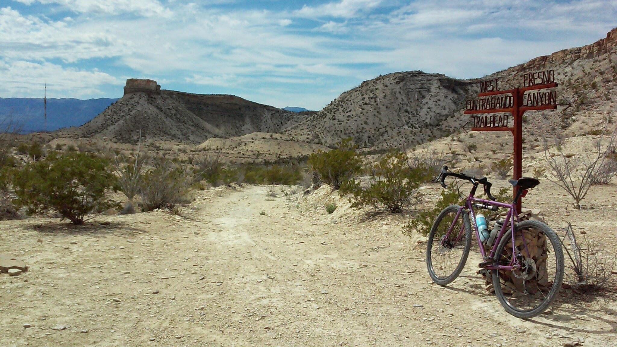 A mountain bike resting on a dirt path next to a wooden signpost indicating directions to various trails, set against a backdrop of dry, rocky hills and a blue sky with scattered clouds. Big Bend Ranch State Park mountain bike trail.