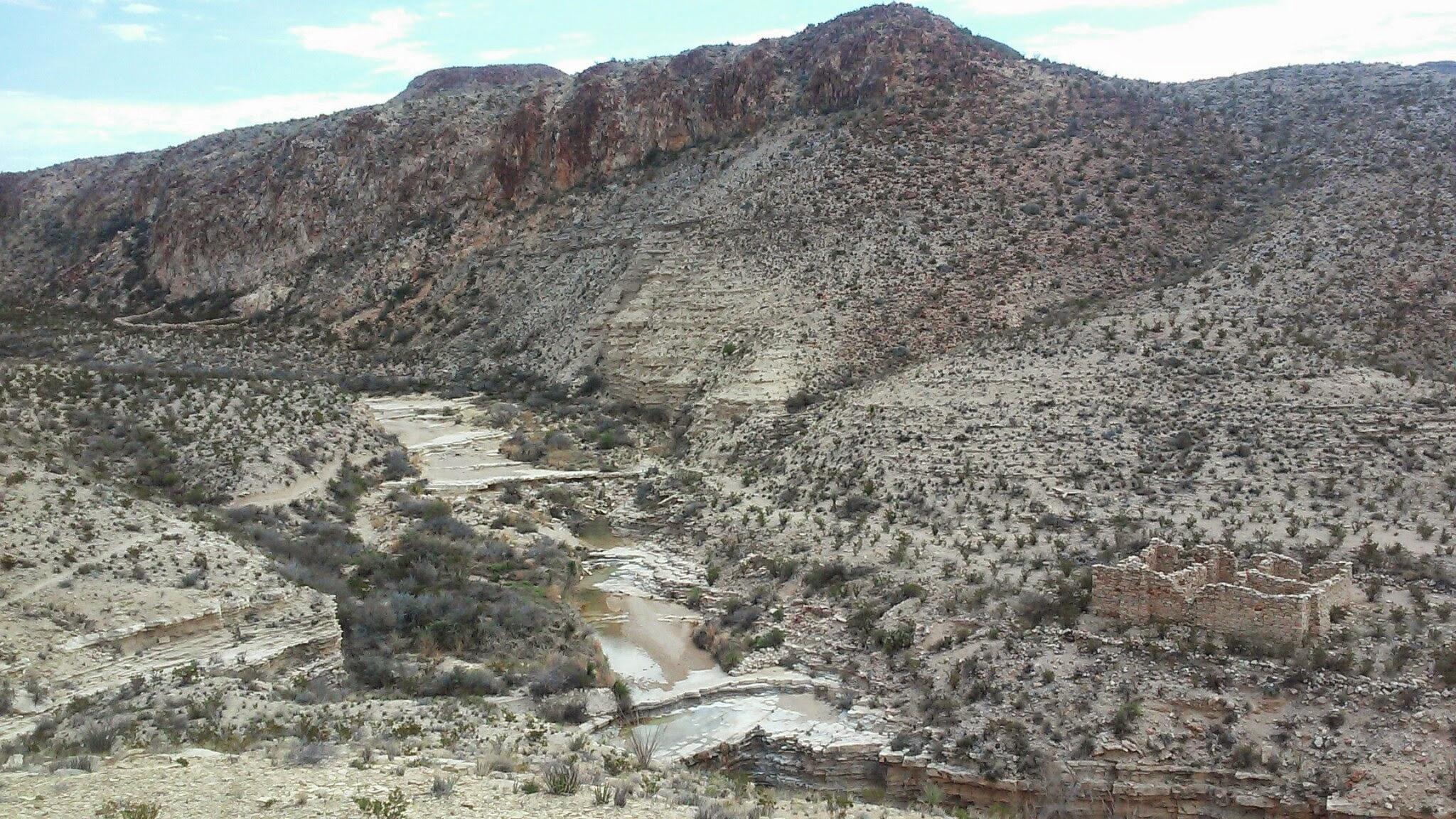 A scenic view of a rocky canyon landscape featuring steep, rugged cliffs and a winding riverbed. Sparse vegetation and scattered shrubs dot the sandy terrain, while remnants of an old stone structure are visible on the right side of the image. The sky above is partly cloudy, adding depth to the arid setting. Big Bend State Park mountain bike trail.