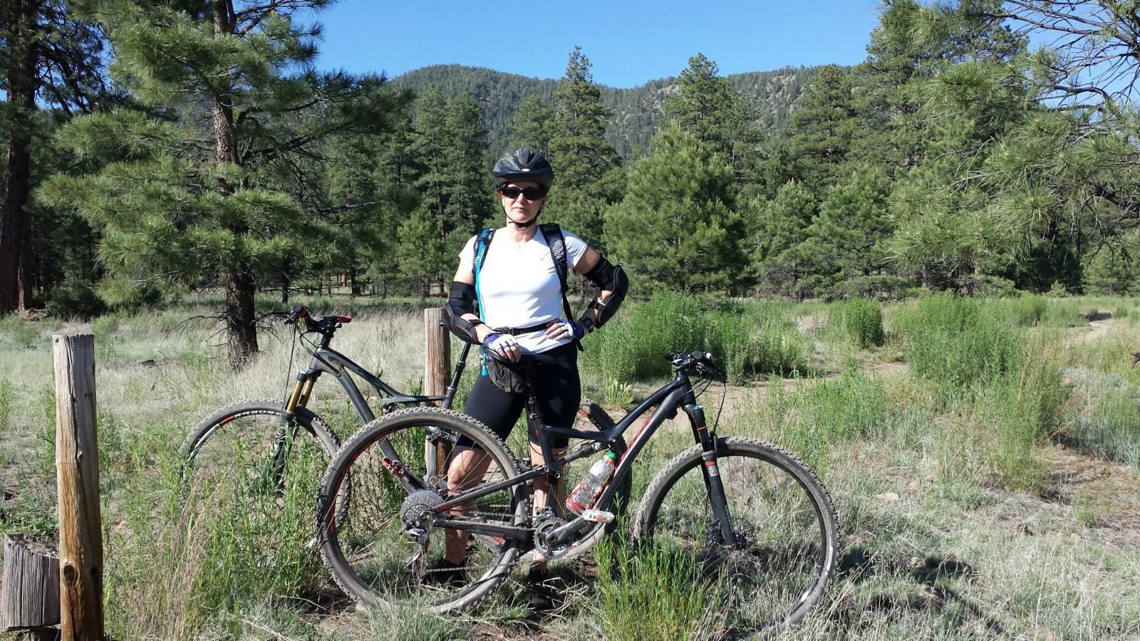 Specialized Rumor: A cyclist wearing a helmet and sunglasses stands confidently beside two mountain bikes in a grassy area surrounded by trees and mountains. The scene is bright and sunny, reflecting a perfect day for outdoor biking adventures.