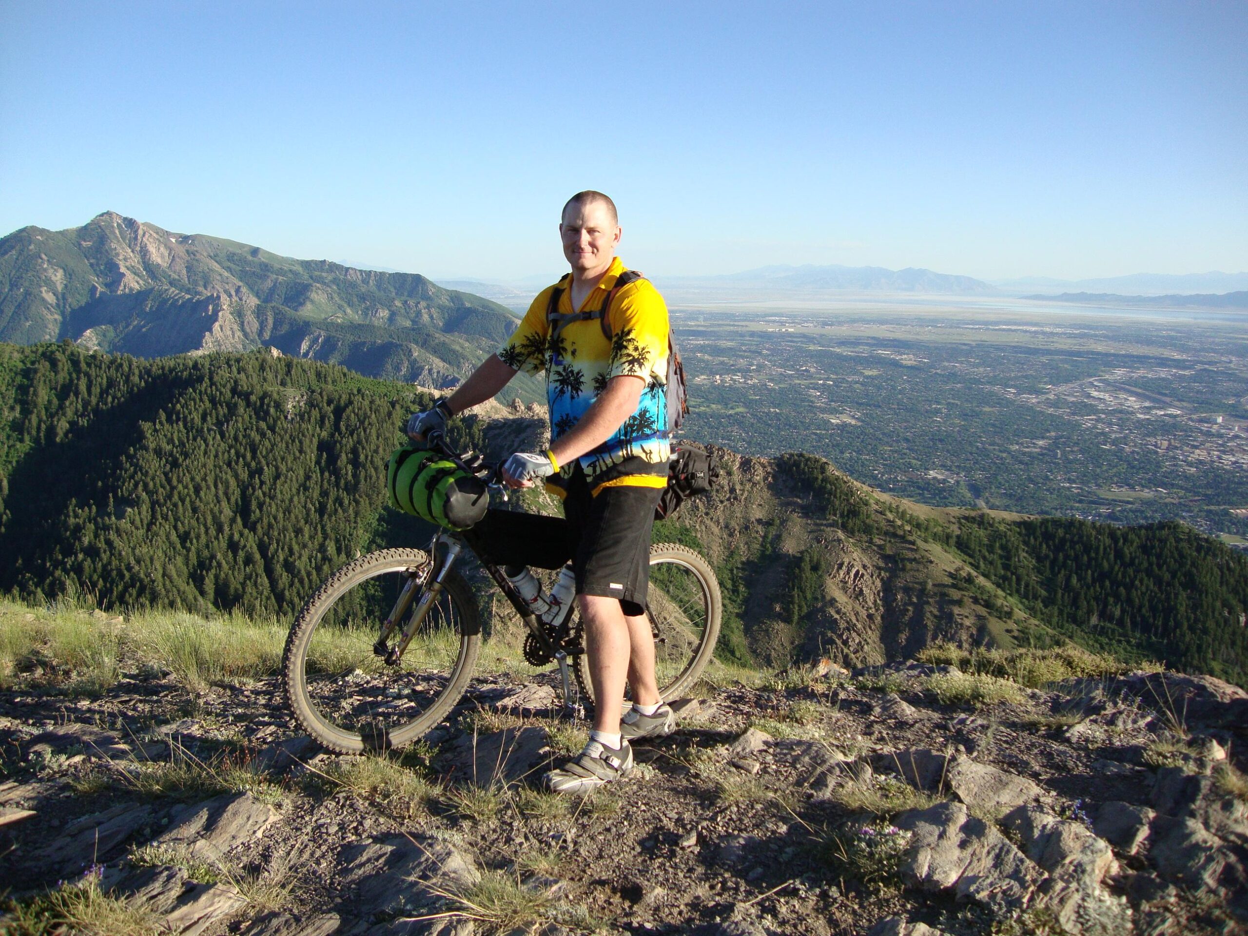 A person stands next to a mountain bike on a rocky summit, overlooking a vast valley and distant mountains. The individual wears a colorful shirt with a tropical pattern and black shorts, and has a camping mat strapped to the bike. The scene is bathed in bright sunlight, showcasing lush green forests and a clear blue sky. Bonneville Shoreline Trail - Ogden Section mountain bike trail.