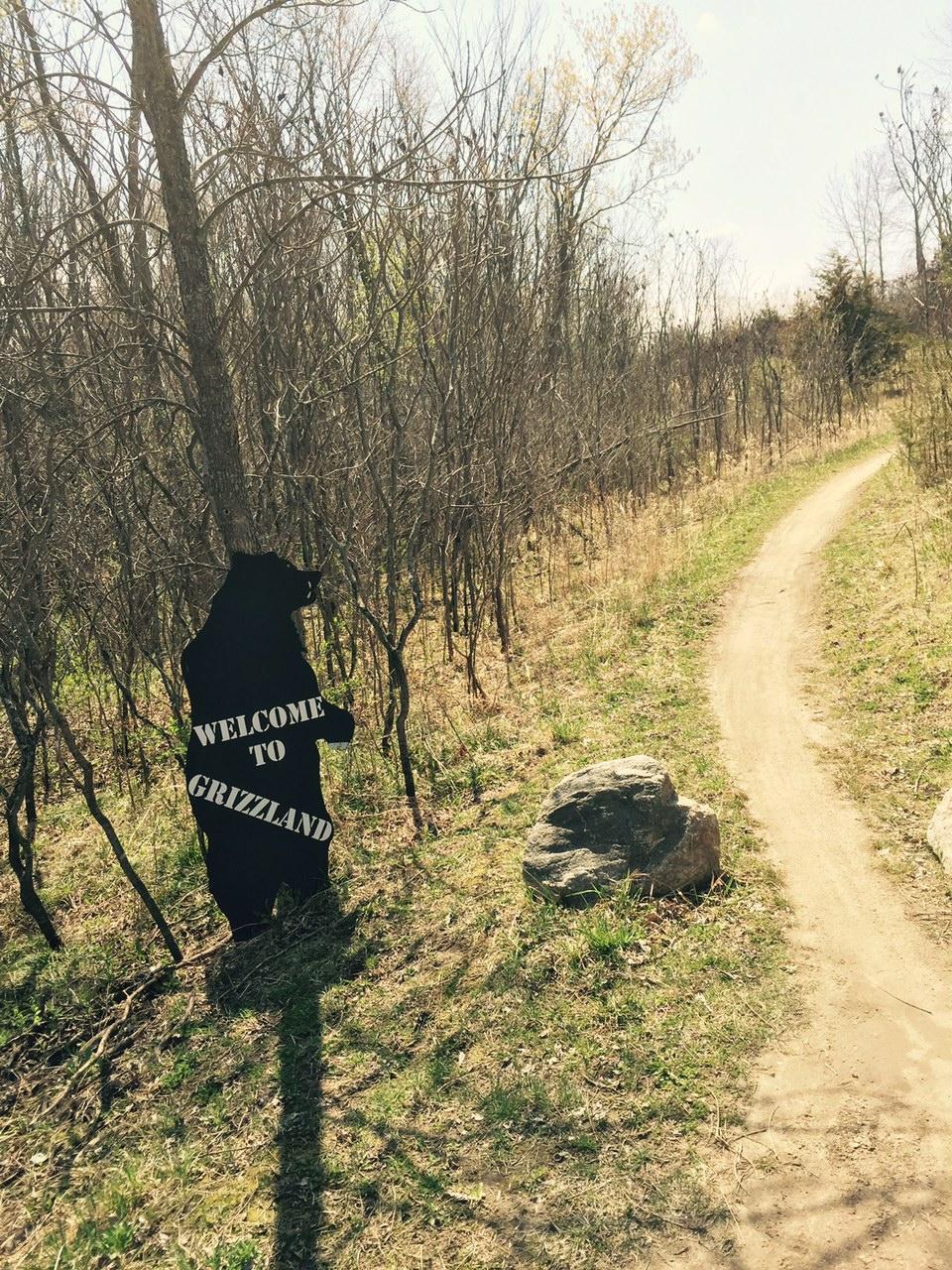 A trail surrounded by bare trees, with a black bear silhouette holding a sign that reads "WELCOME TO GRIZZLAND," positioned near a dirt path. A large rock is visible on the ground beside the trail. Elm Creek Park mountain bike trail.