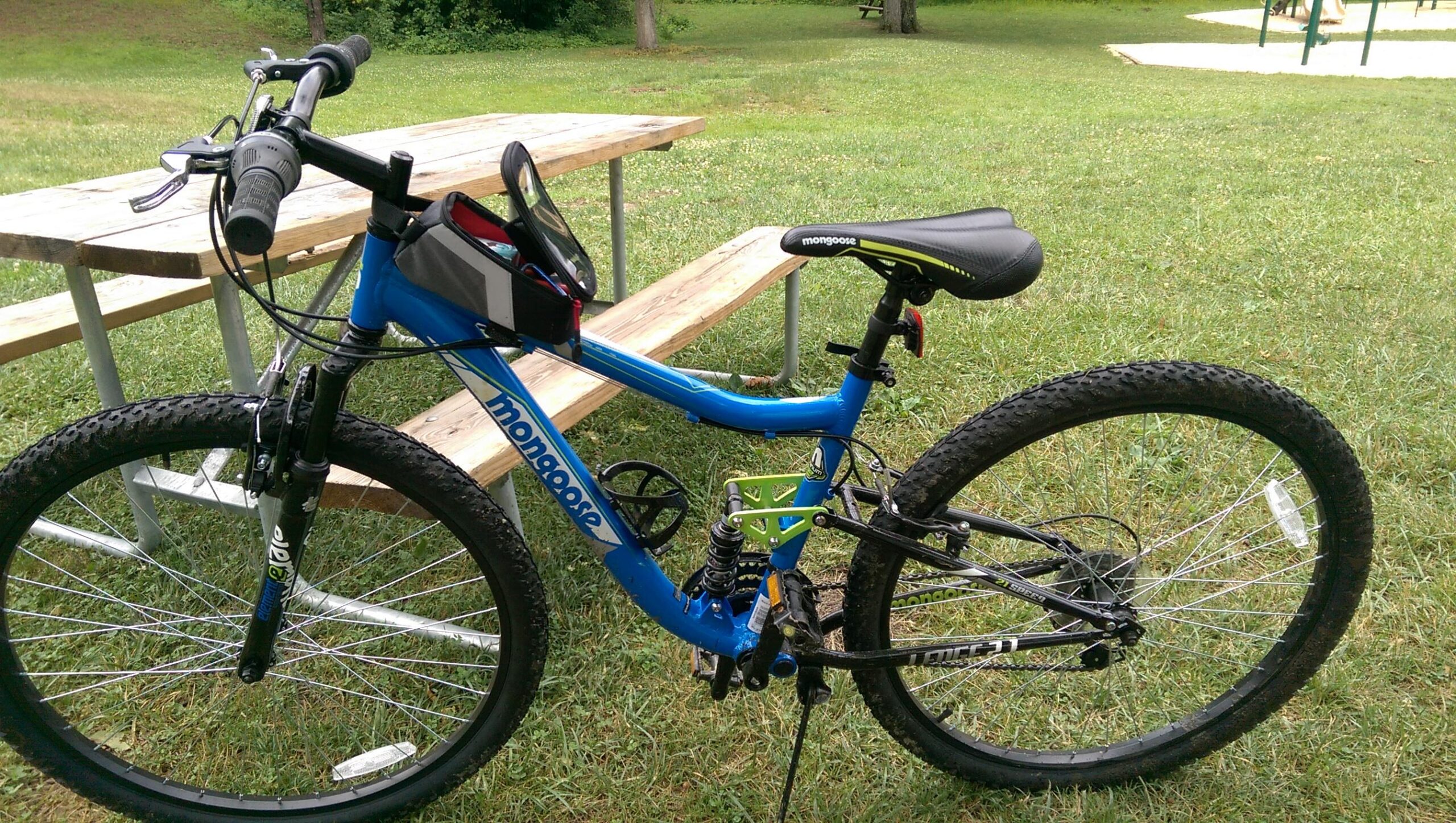Mongoose Mongoose Ledge 2.1: A blue Mongoose mountain bike parked on grass near wooden picnic tables. The bike features thick tires, a suspension system, and a black seat with green accents. In the background, there is a playground visible.