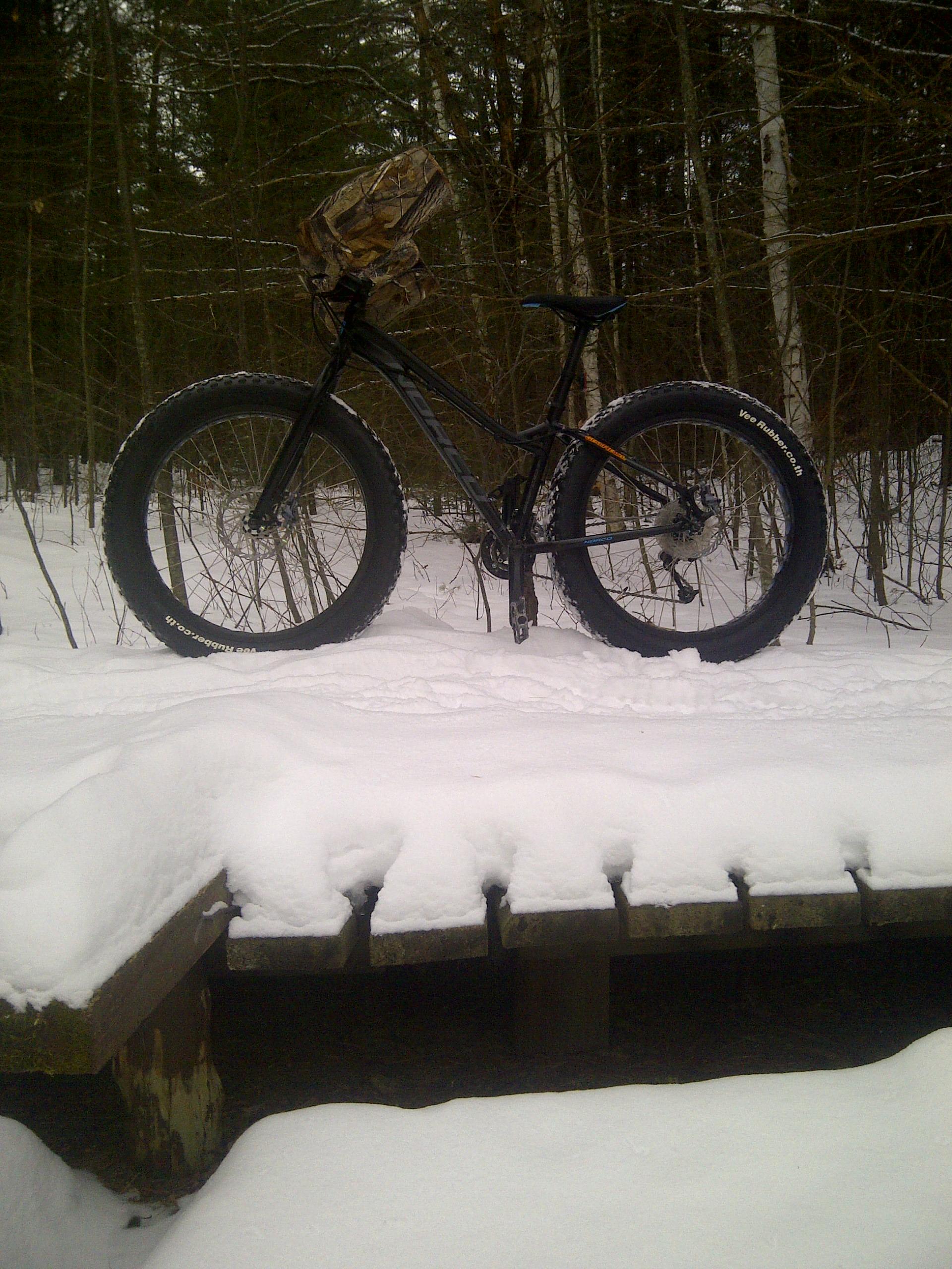 Norco Bigfoot: A fat tire bike standing on a wooden platform covered in snow, surrounded by a forest of trees in winter. The bike features large, wide tires suitable for snow, and has a camouflage backpack attached to the handlebars.