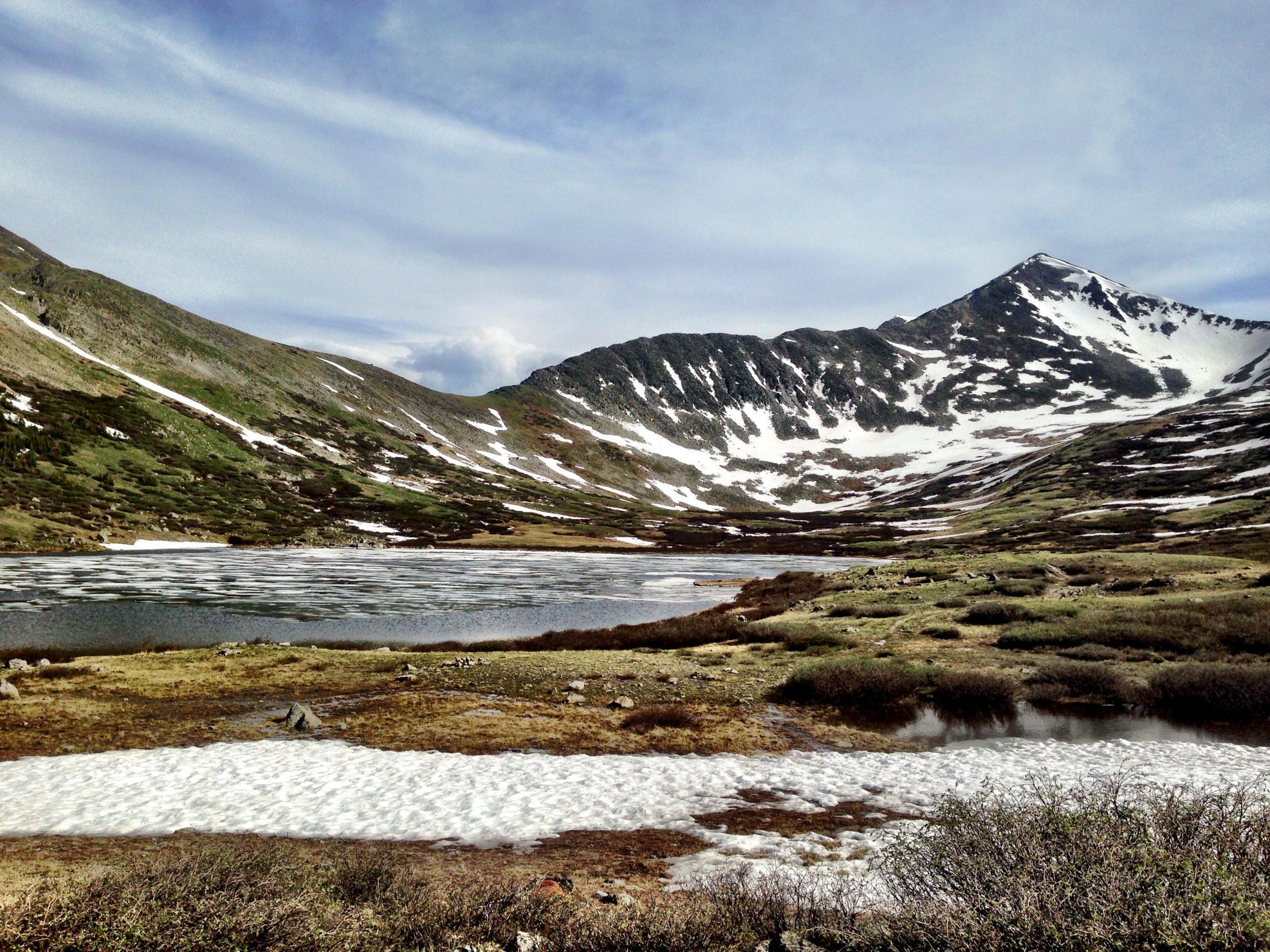 A scenic mountain landscape featuring a tranquil lake surrounded by snow-capped peaks and green rocky slopes. The foreground includes patches of melting snow and diverse vegetation under a partly cloudy sky. CDT: Hancock Lakes mountain bike trail.