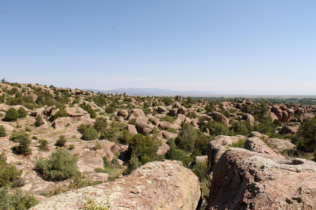 A panoramic view of a rocky landscape filled with large boulders and greenery under a clear blue sky. The terrain features scattered shrubs and small trees, with distant mountains visible on the horizon. Stone Quarry mountain bike trail.