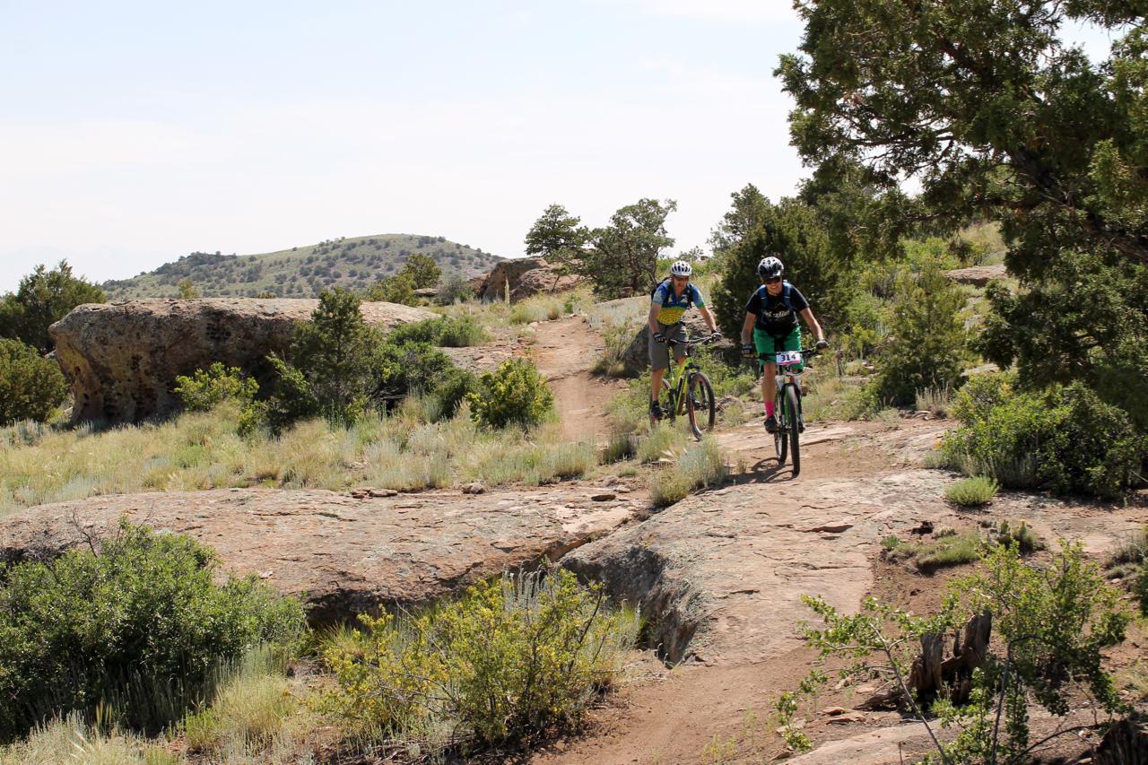 Two mountain bikers navigate a rocky trail through a scenic outdoor landscape. The trail is surrounded by shrubs and small trees, with rolling hills visible in the background under a clear sky. One cyclist is wearing a blue and yellow jersey, while the other is seen in a black jersey and pink shorts. Stone Quarry mountain bike trail.