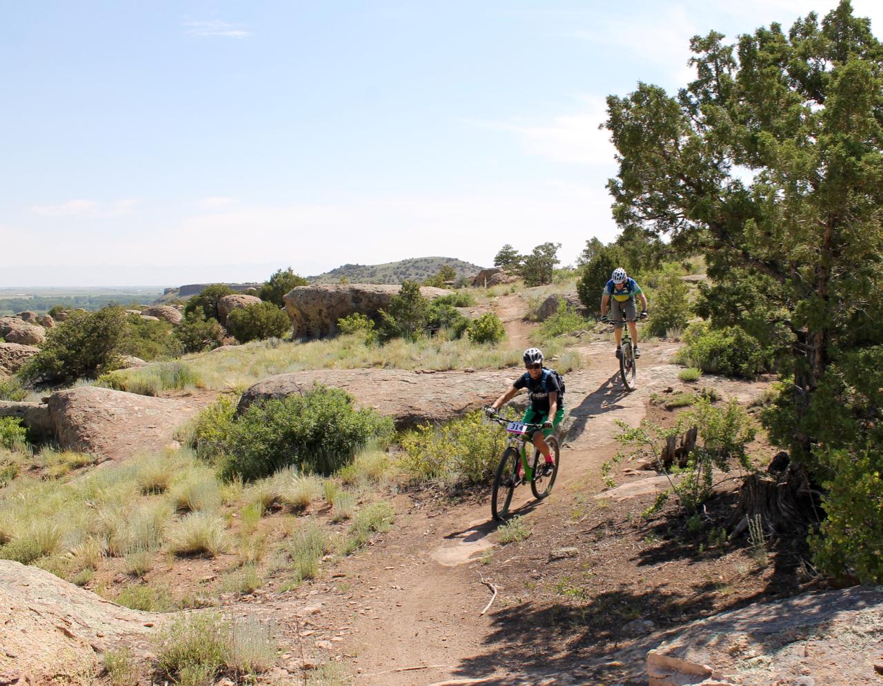 Two mountain bikers navigate a rocky trail surrounded by greenery and shrubs under a clear blue sky. The landscape features large boulders and rolling hills in the background, highlighting an outdoor adventure setting. Stone Quarry mountain bike trail.