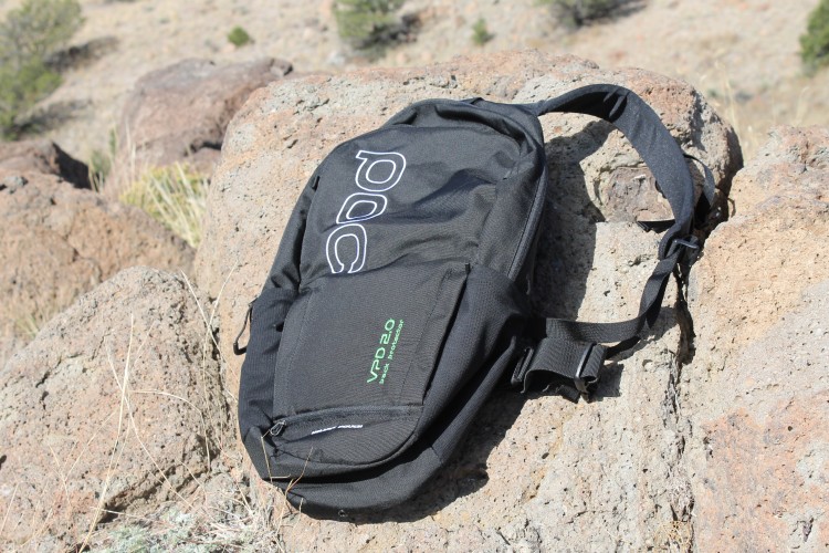 A black backpack with the logo "POC" and "VPD 20" on it, resting on a rugged stone surface in a natural outdoor setting with sparse vegetation and rocky terrain.