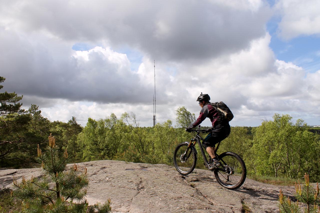 A mountain biker rides on a rocky outcrop, overlooking a lush green landscape with trees. In the background, a tall communication tower rises against a cloudy sky. Skatas Trail System mountain bike trail.
