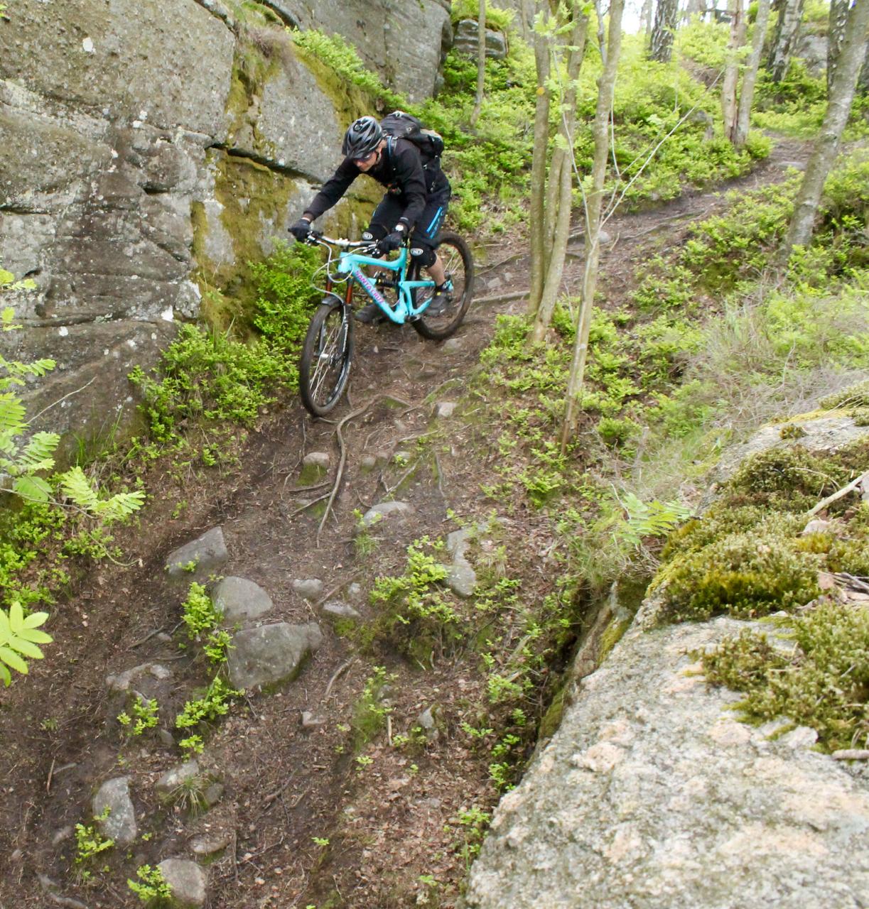 A mountain biker navigates a narrow, rocky trail surrounded by lush greenery and towering rocks. The cyclist leans forward while pedaling, demonstrating skill and balance on the uneven terrain. Skatas Trail System mountain bike trail.