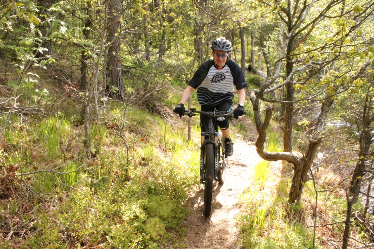 A person riding a mountain bike along a narrow trail in a lush green forest. The cyclist is wearing a helmet and sunglasses, and is surrounded by trees and greenery. Sunlight filters through the leaves, illuminating the path. Kasjon Lake Trails mountain bike trail.